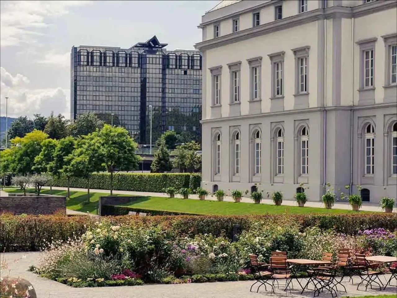 Blick über eine Parkanlage mit Blumenbeeten und Café-Tischen in Koblenz, rechts ein historischer Gebäudeflügel, im Hintergrund ein modernes Glasgebäude