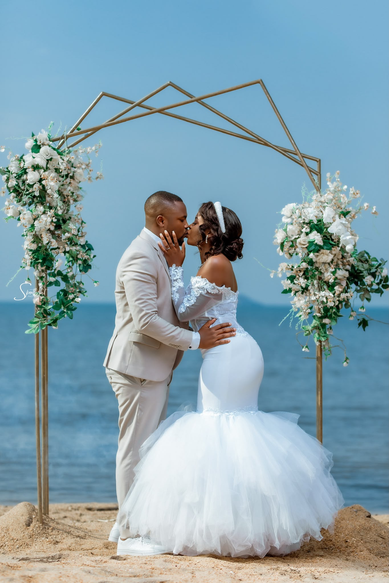 A couple kissing beneath a gold geometric arch on the beach at Senga Bay