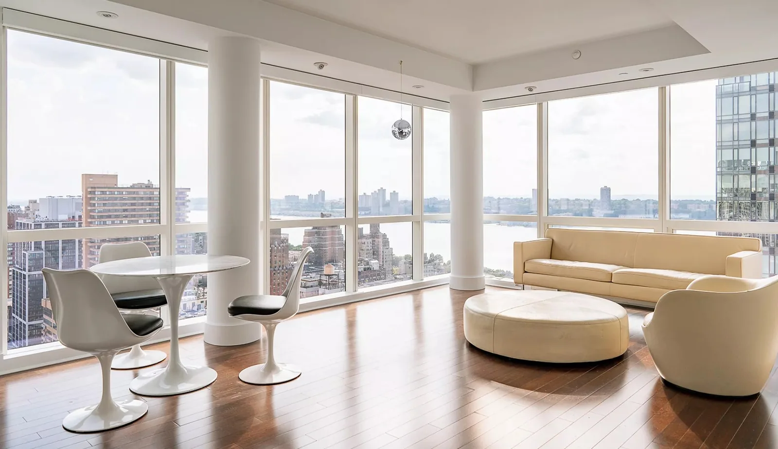 Sunlit New York City apartment living room with floor-to-ceiling windows, a cream sofa, and a round dining table.