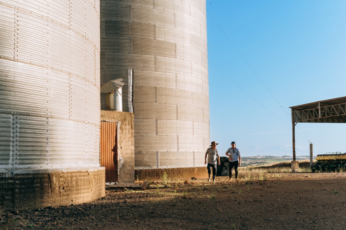 Two people walking past large grain silos on a sunny day
