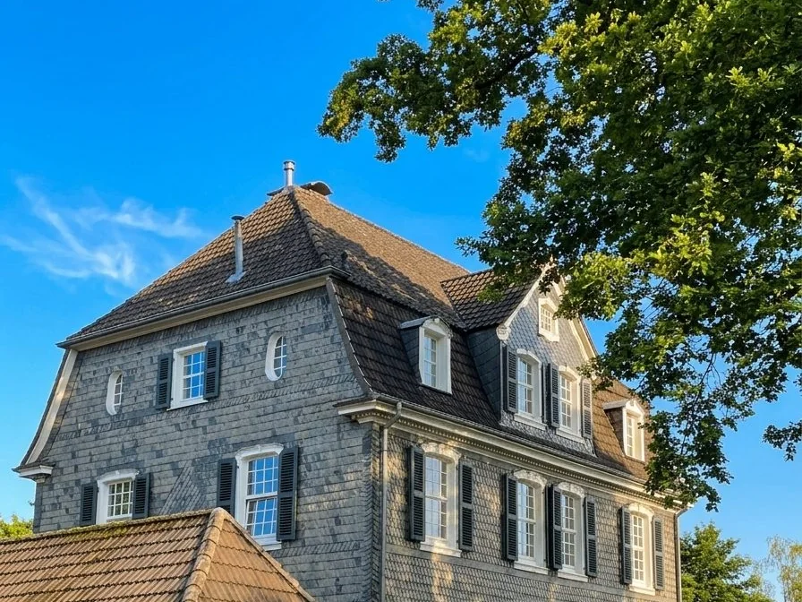 Altes Schieferhaus mit dunklen Fensterläden in Bergisch Gladbach, davor ein großer Baum