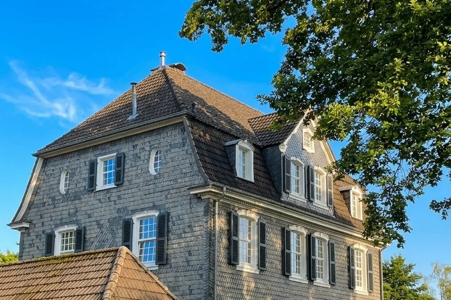 Altes Schieferhaus mit dunklen Fensterläden in Bergisch Gladbach, davor ein großer Baum