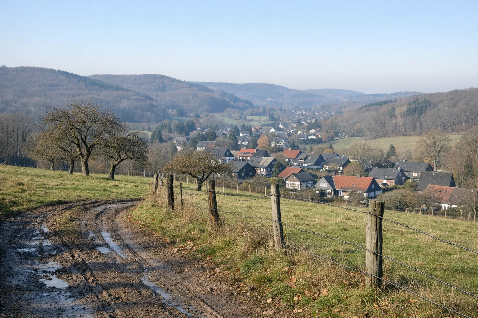 Feldweg mit Weidezaun und alten Obstbäumen über einem Dorf im Tal, dahinter bewaldete Hügel