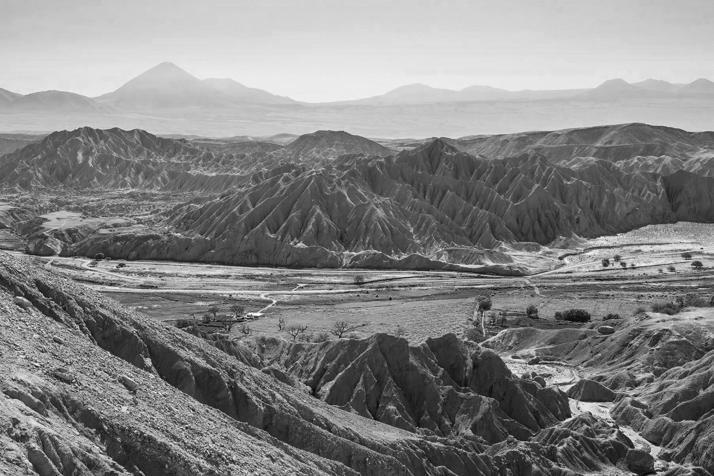Eroded ridges of the Cordillera de la Sal above a green valley floor, with volcanoes on the horizon, in black and white