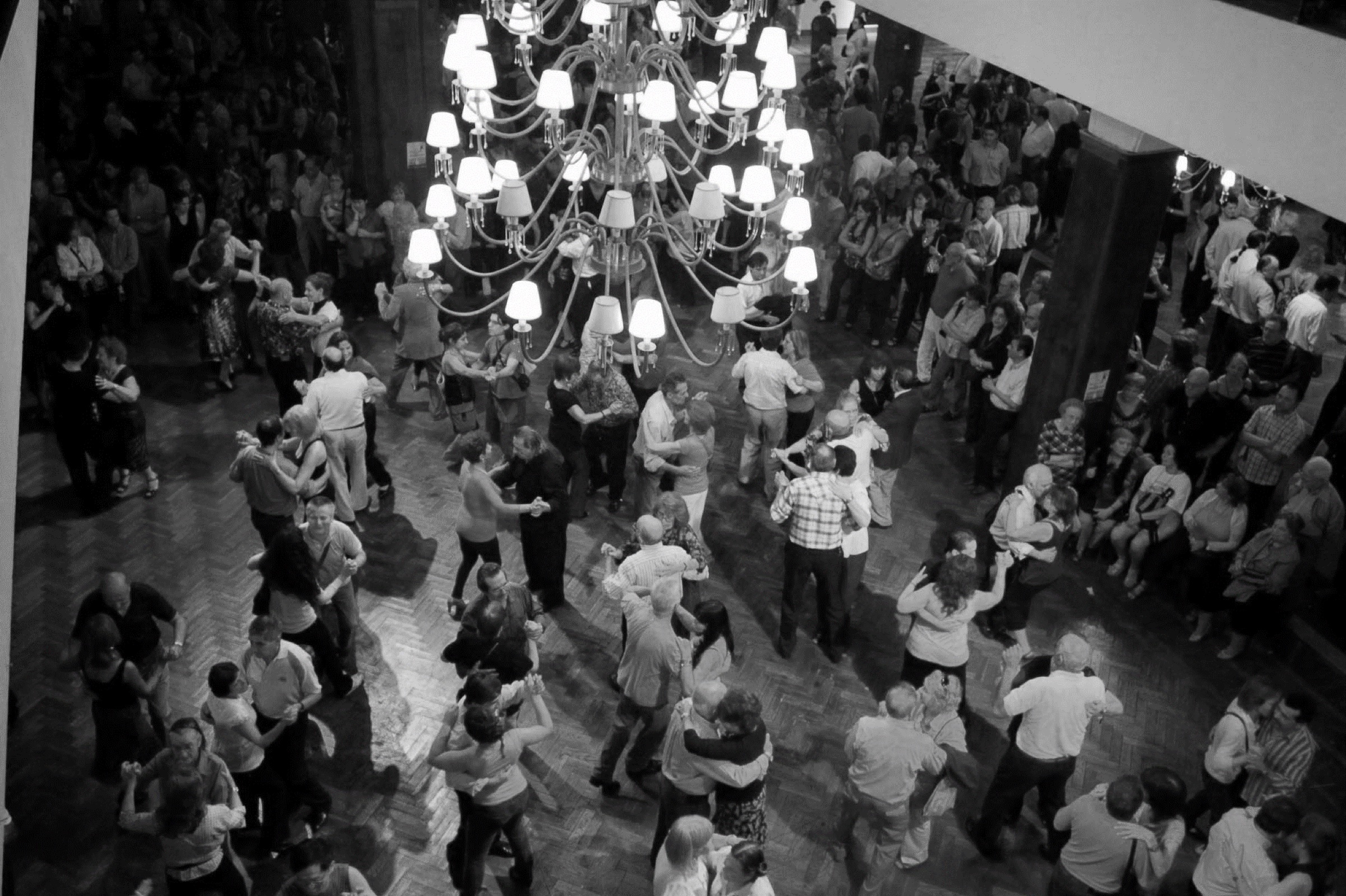 Couples dancing tango on a crowded milonga floor under a large chandelier, seen from above