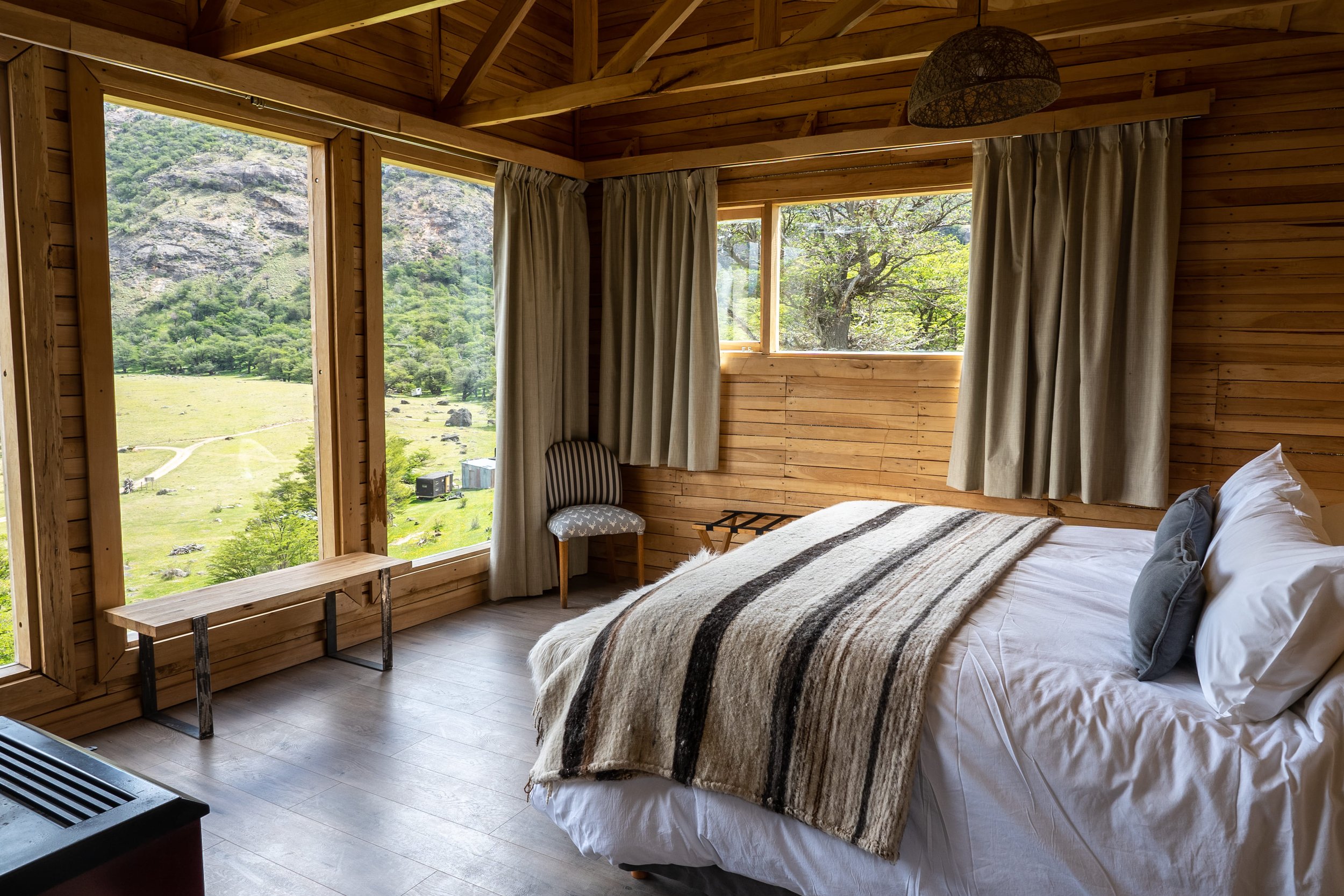 Habitación de madera con cama doble y grandes ventanales con vista a las montañas y valles.