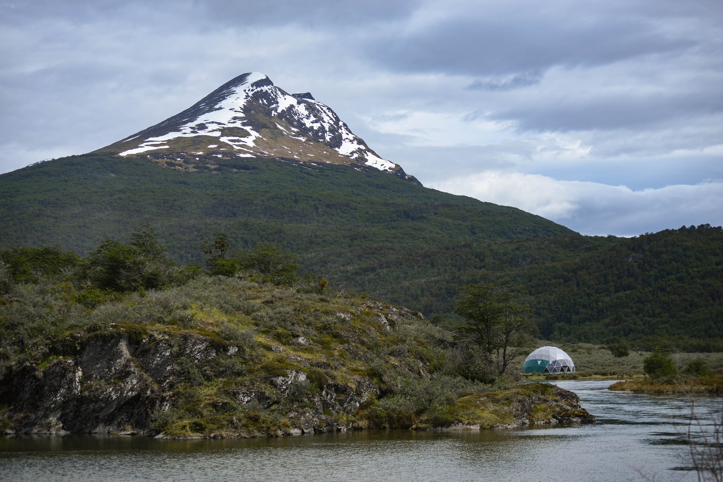 Domo geodésico a orillas del agua con una montaña de cumbre nevada y bosque de fondo.