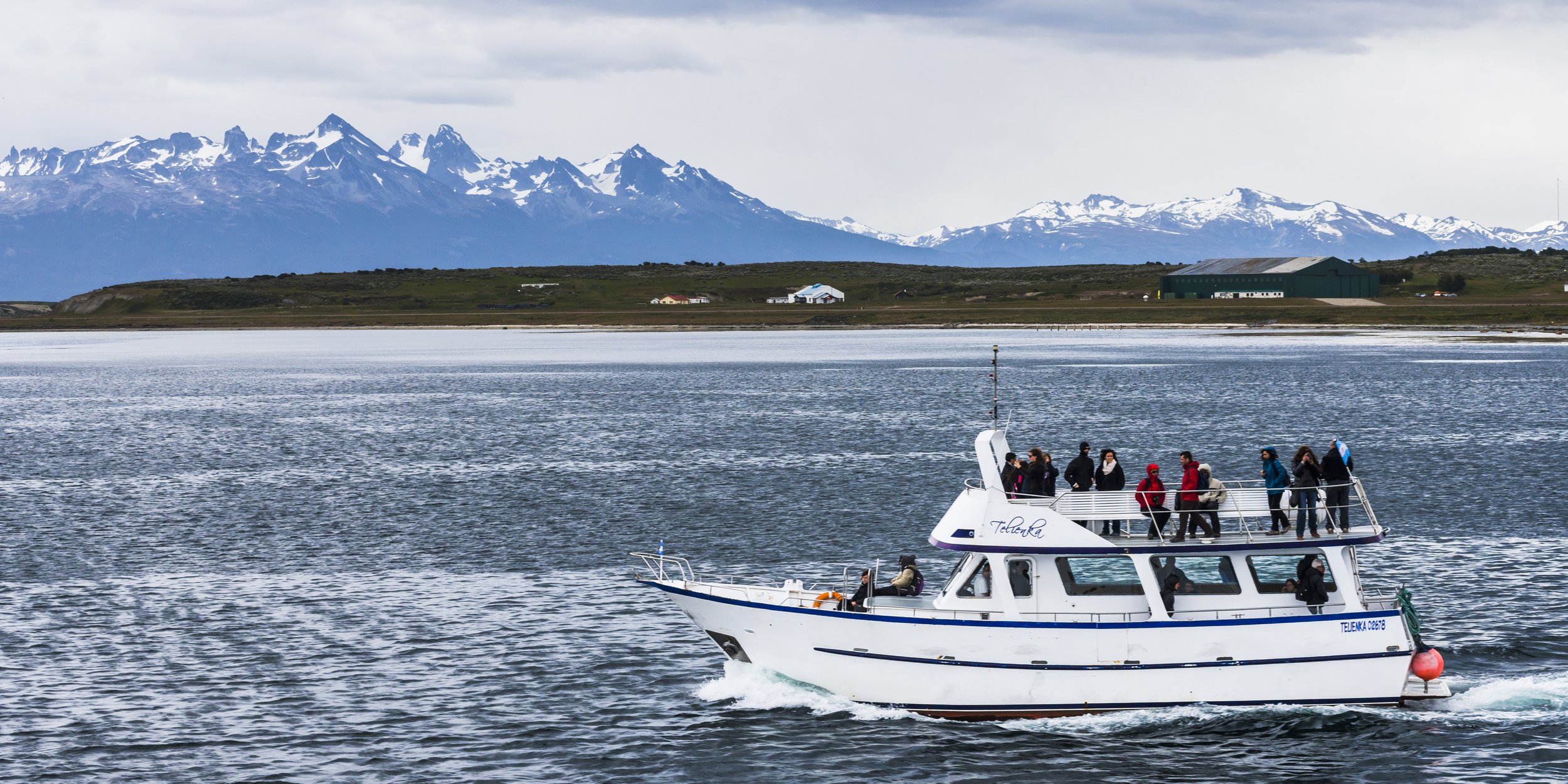Barco turístico navegando por el Canal Beagle con pasajeros en cubierta y montañas nevadas de fondo.