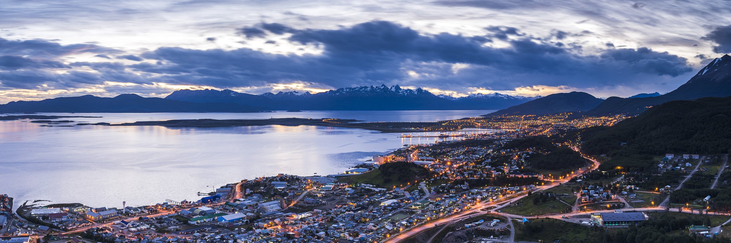 Vista panorámica de Ushuaia iluminada al atardecer frente a la costa del canal Beagle y montañas de fondo.
