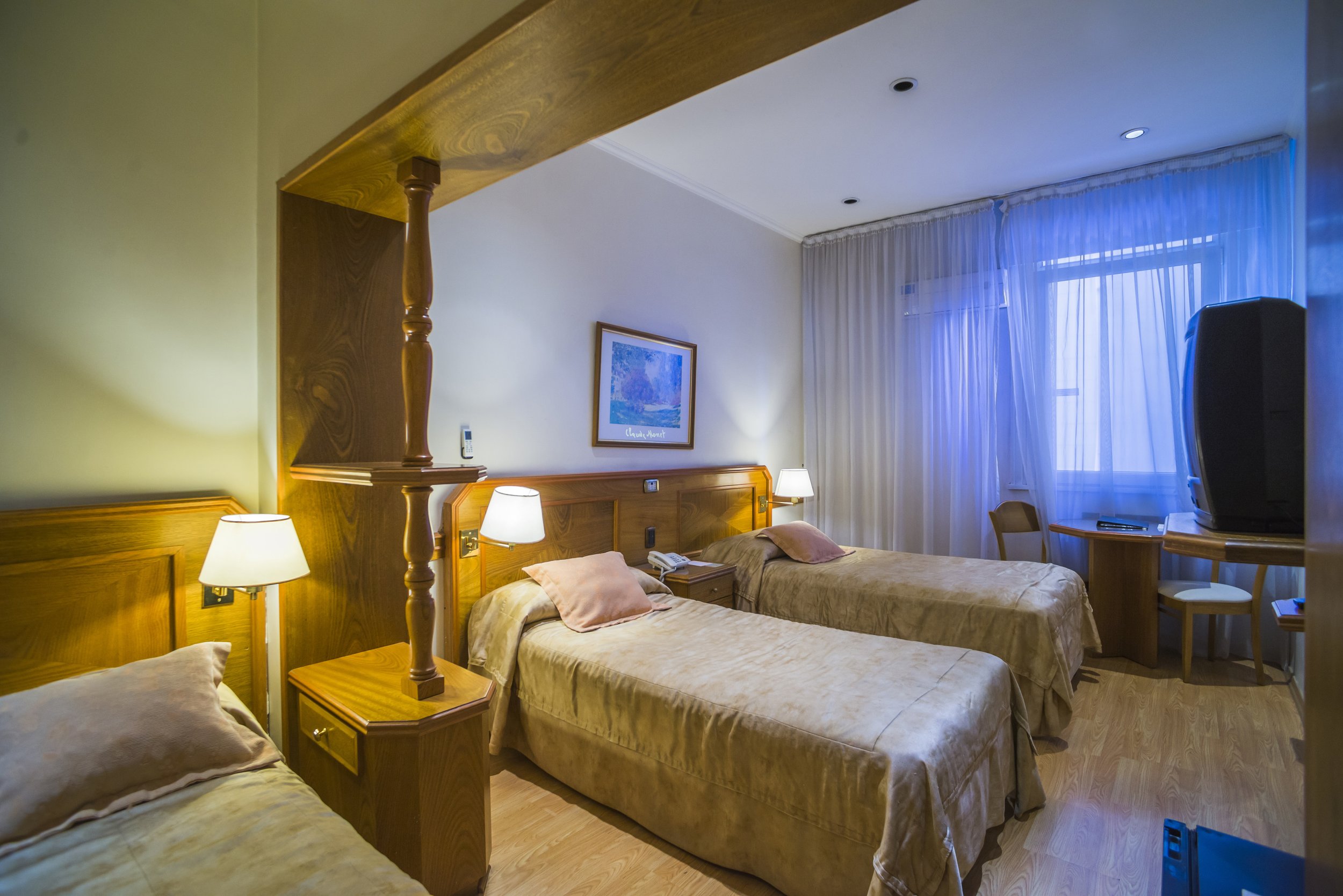 Habitación de hotel con camas individuales, muebles de madera, lámparas de noche y ventana con cortinas.