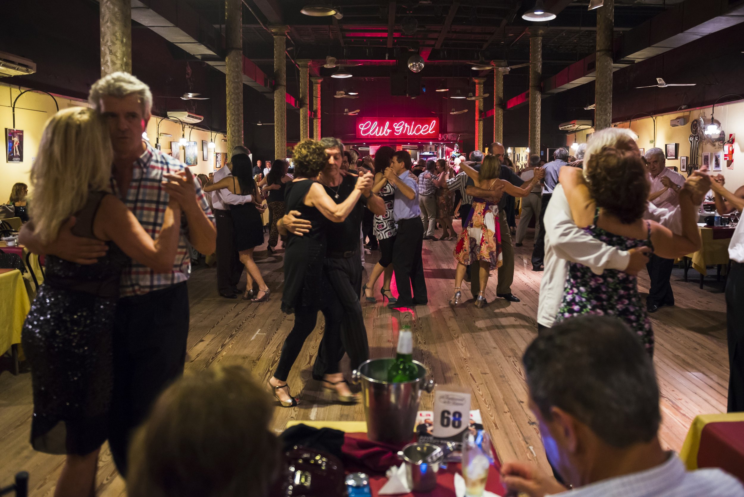 Parejas bailando tango en la pista de madera de una tradicional milonga con mesas alrededor.