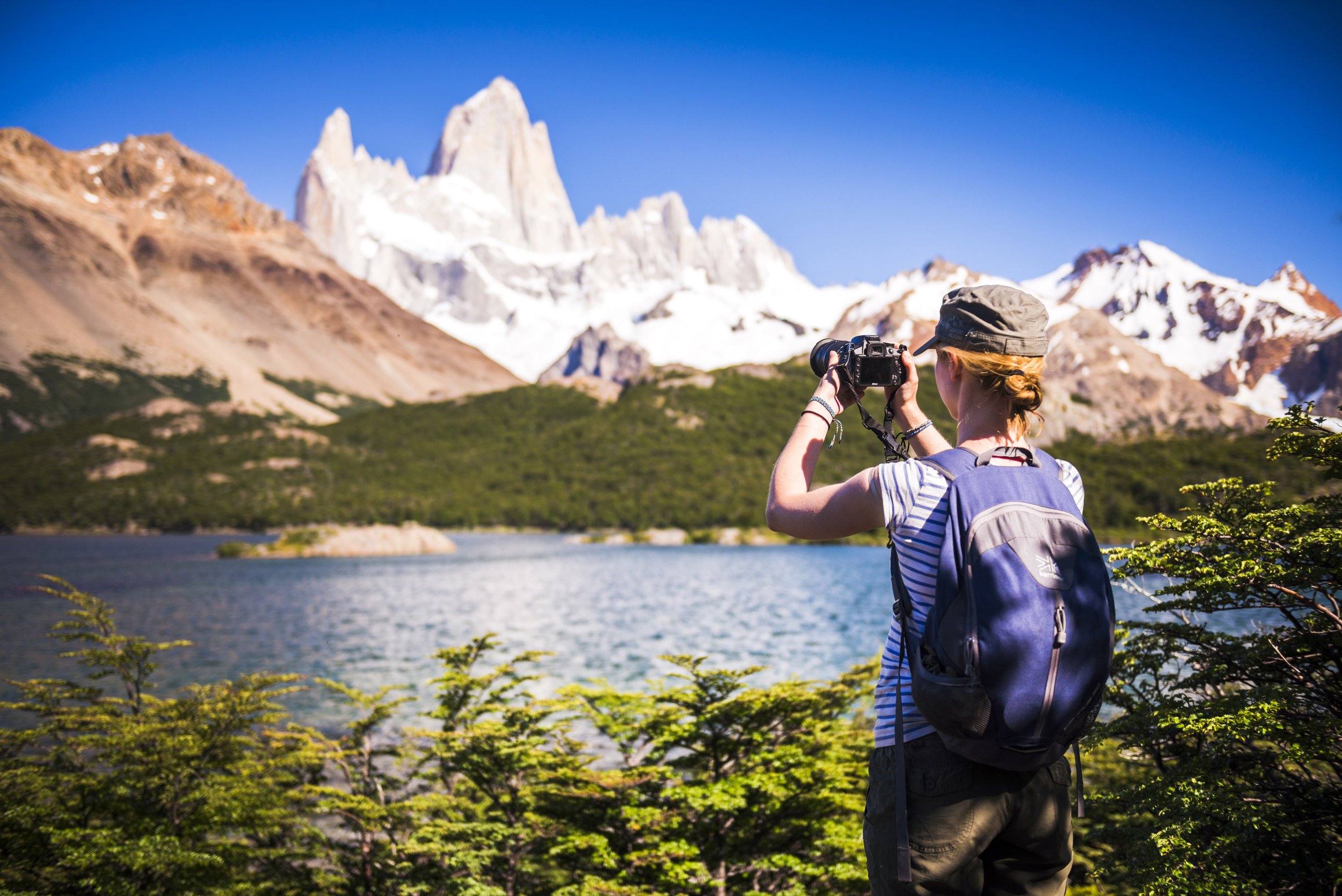 Una mujer de espaldas toma una fotografía frente a la Laguna Capri y el Cerro Fitz Roy nevado.