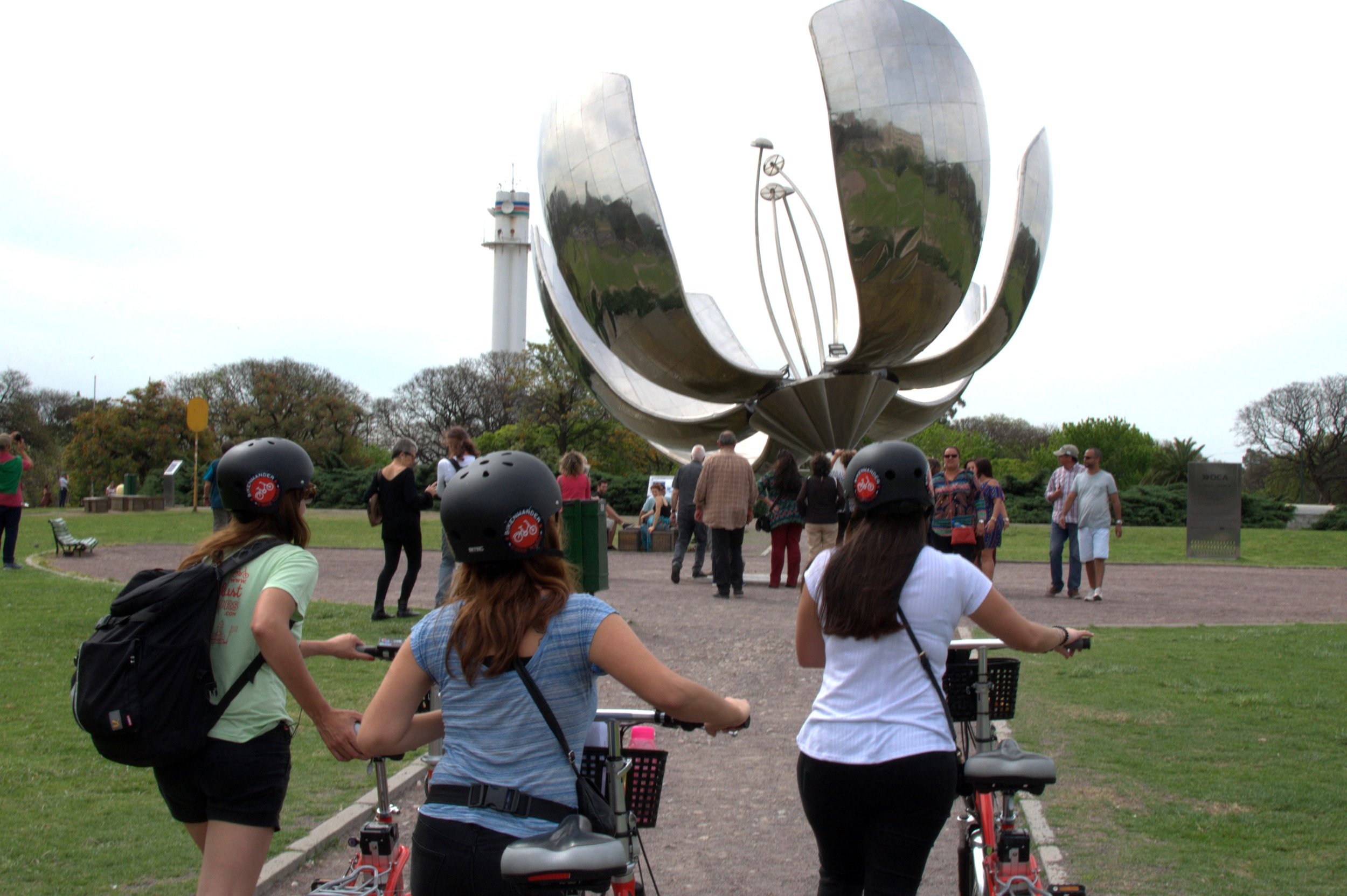 Personas en bicicleta de espaldas observando la escultura metálica Floralis Genérica en una plaza de Buenos Aires.