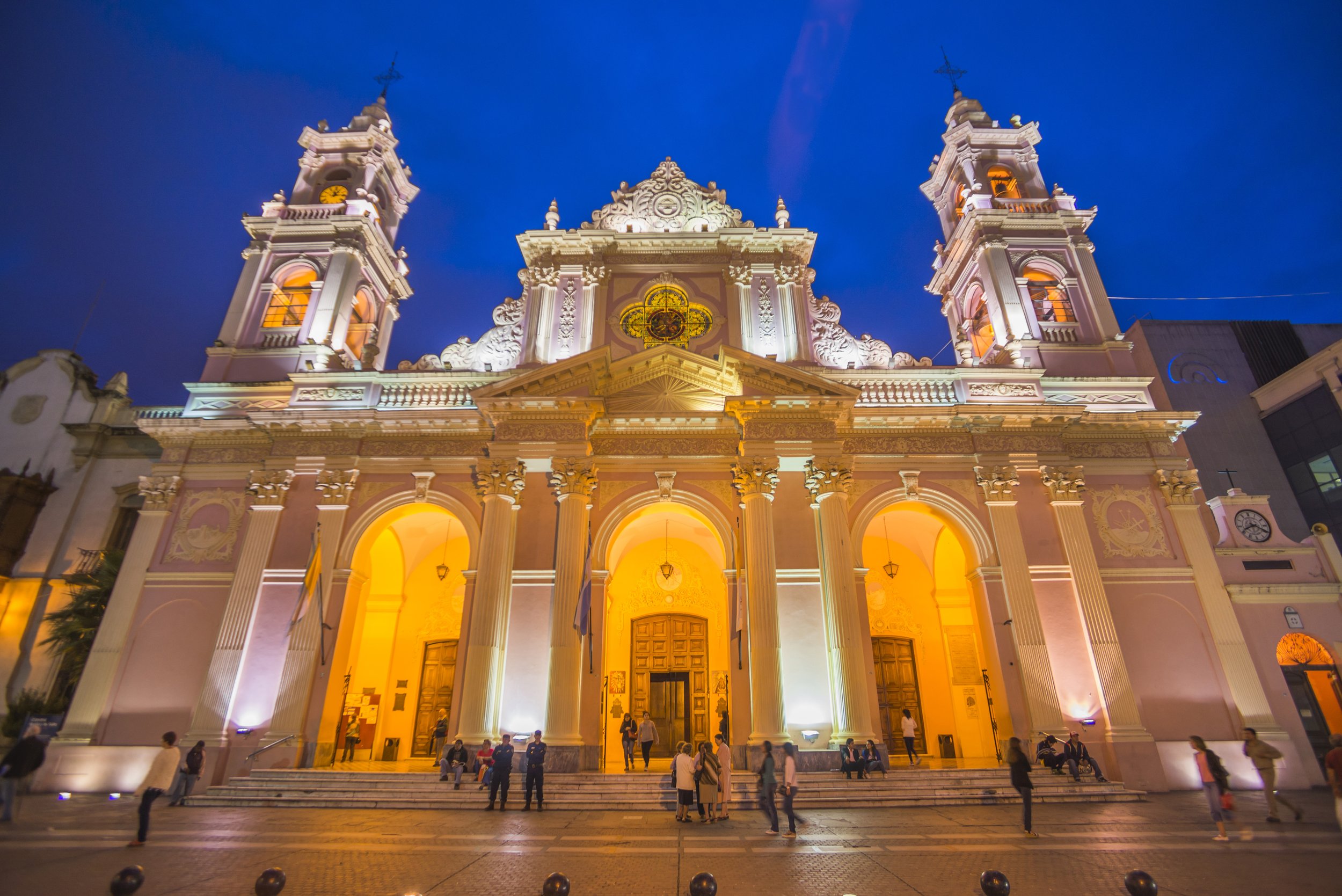 Fachada iluminada de la Catedral Basílica de Salta de noche con personas en la escalinata exterior.
