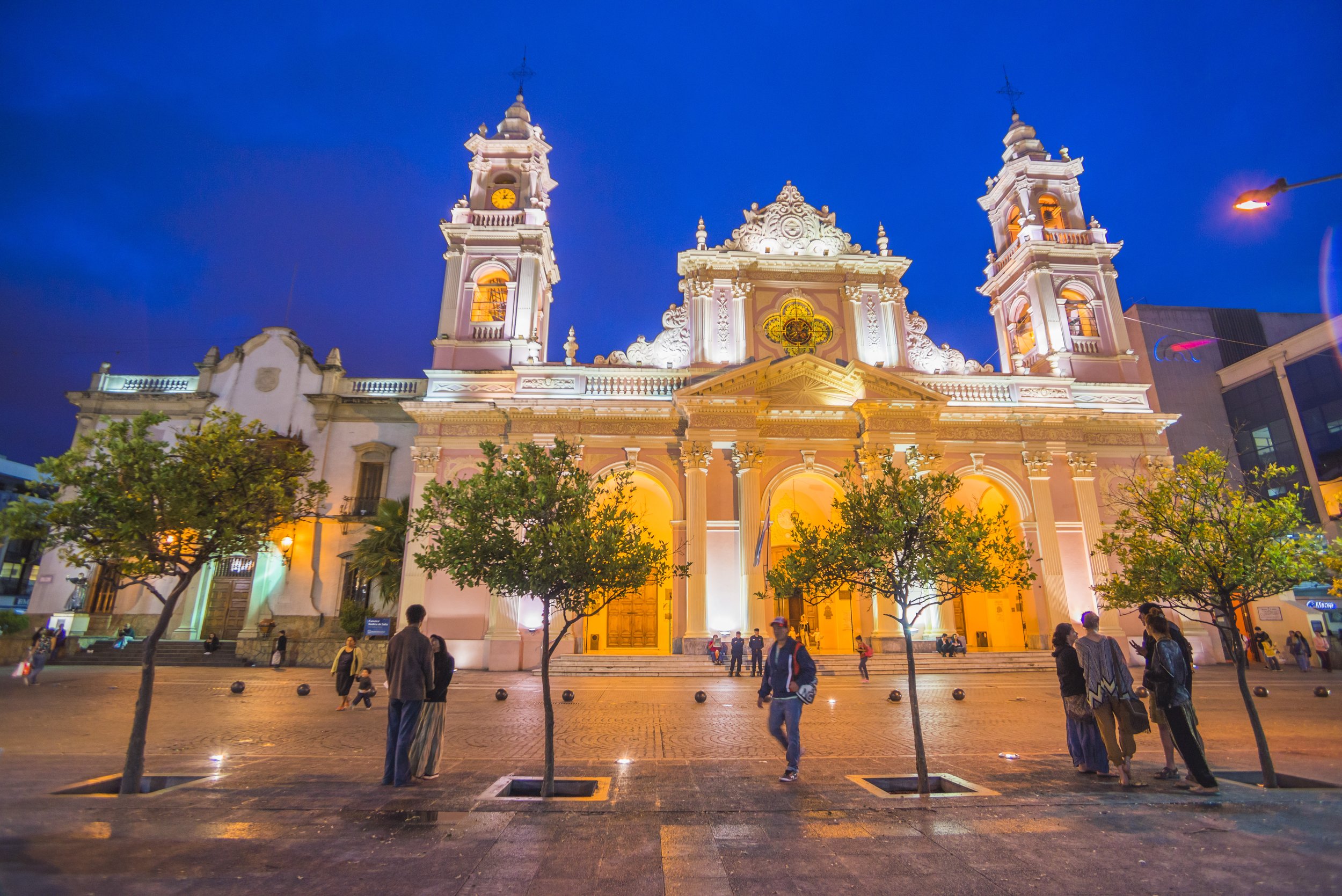 Fachada iluminada de la Catedral de Salta frente a la plaza principal durante la noche con transeúntes.