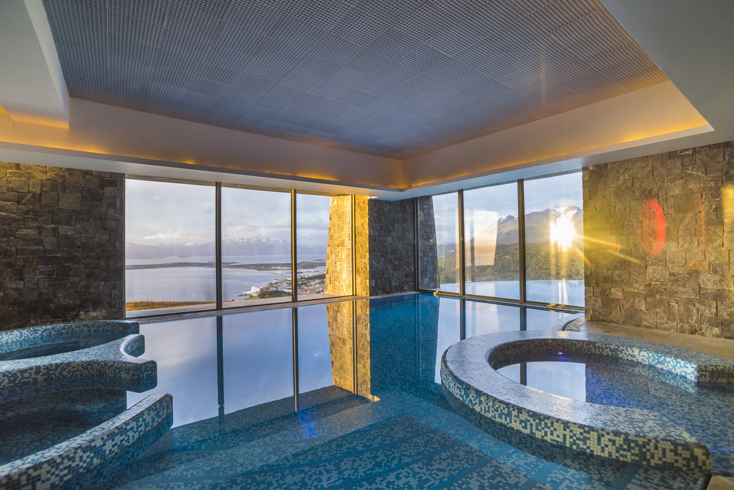 Piscina climatizada e hidromasaje interior con ventanales y vistas panorámicas a la bahía y montañas al atardecer.