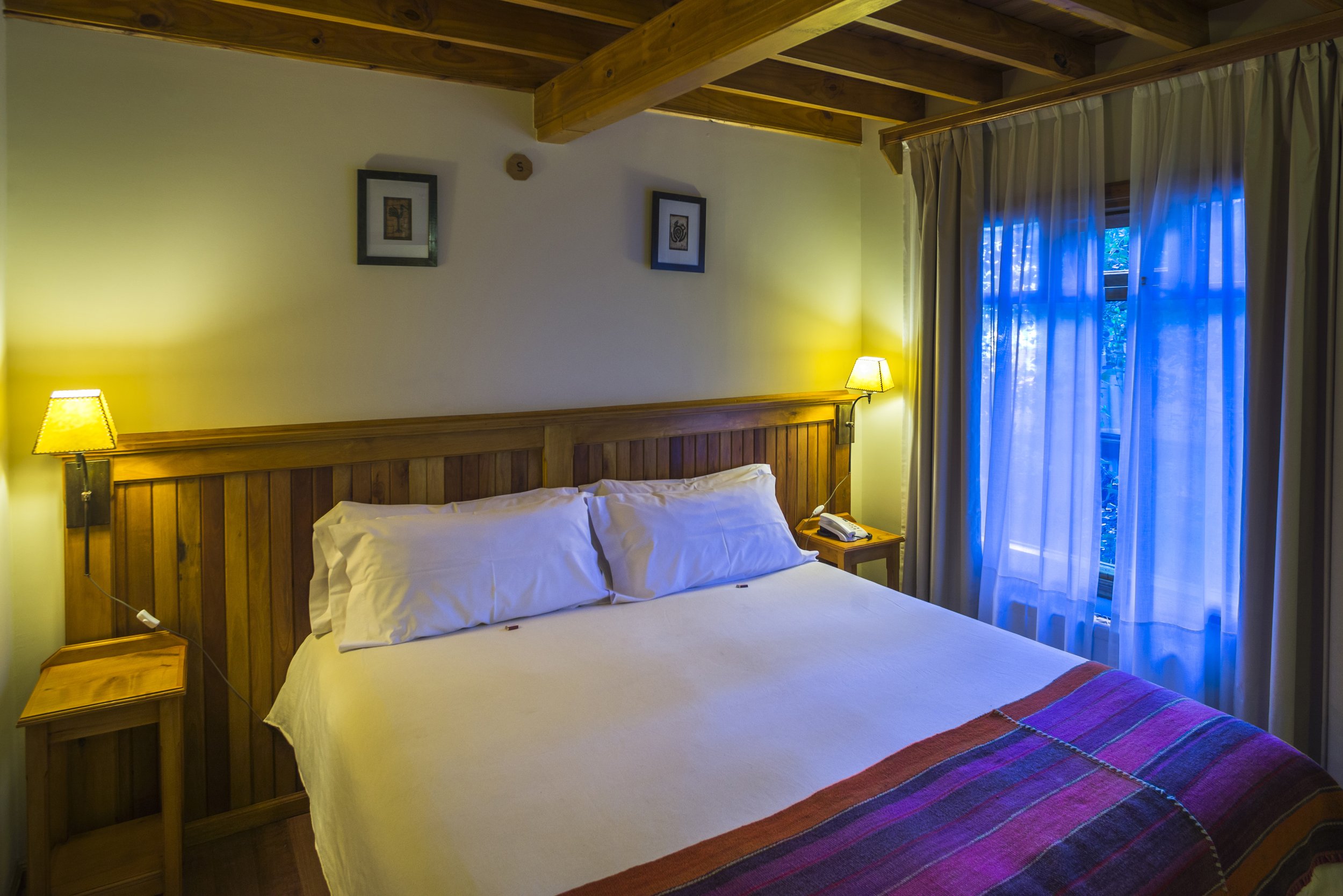 Habitación de hotel con cama doble, respaldo y techo de vigas de madera, y ventana con cortinas.