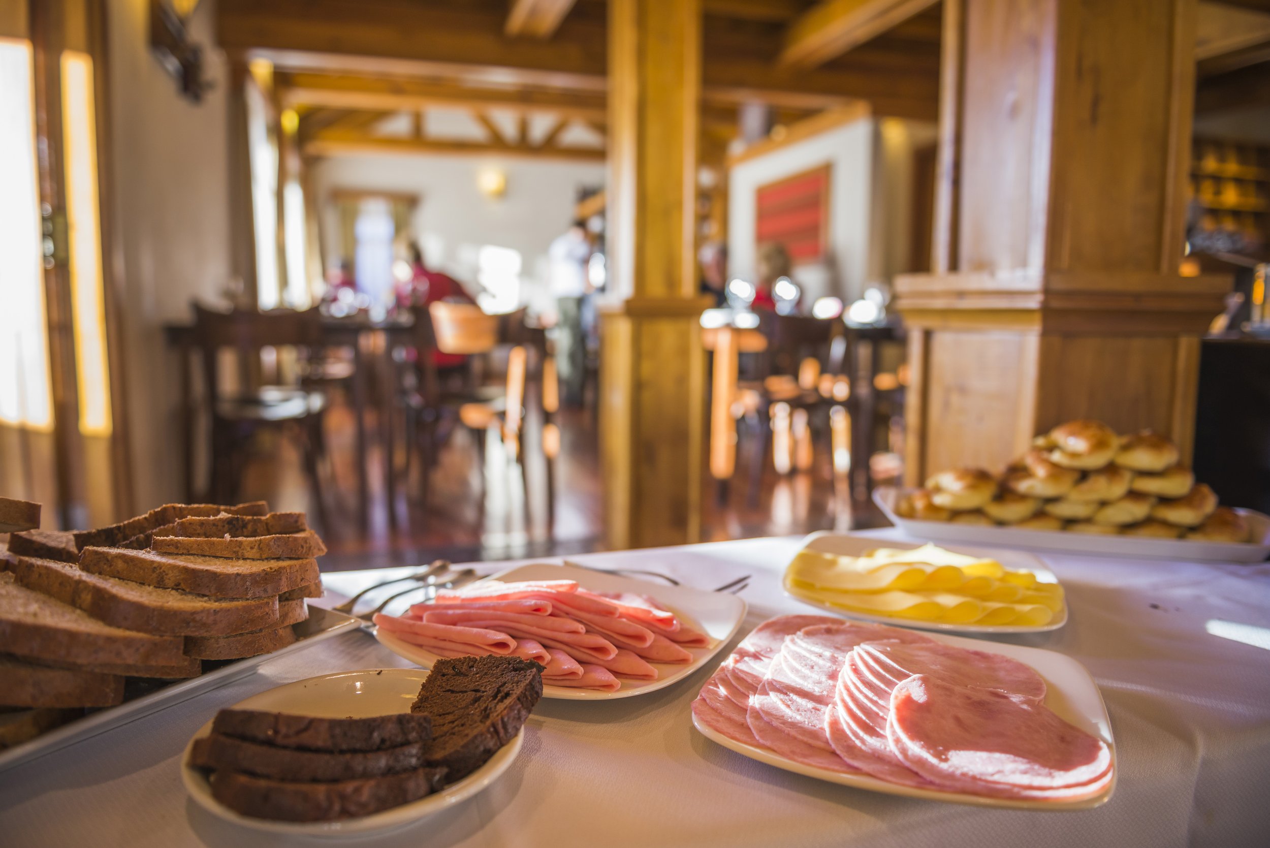 Mesa con fiambres, quesos y panes para el desayuno en el comedor de una hostería con detalles de madera.
