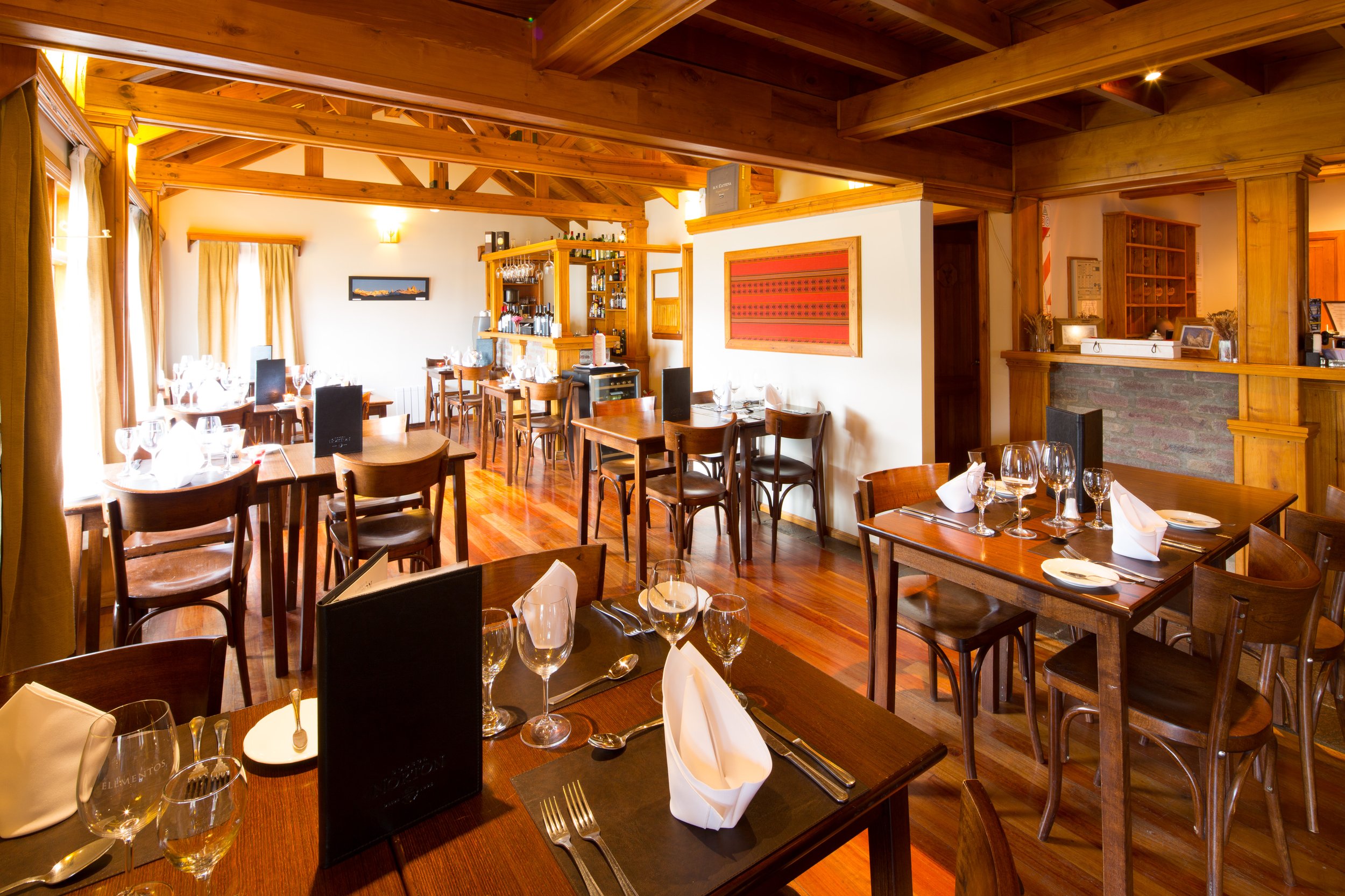 Salón comedor de una hostería con mesas preparadas, sillas de madera y vigas a la vista en el techo.