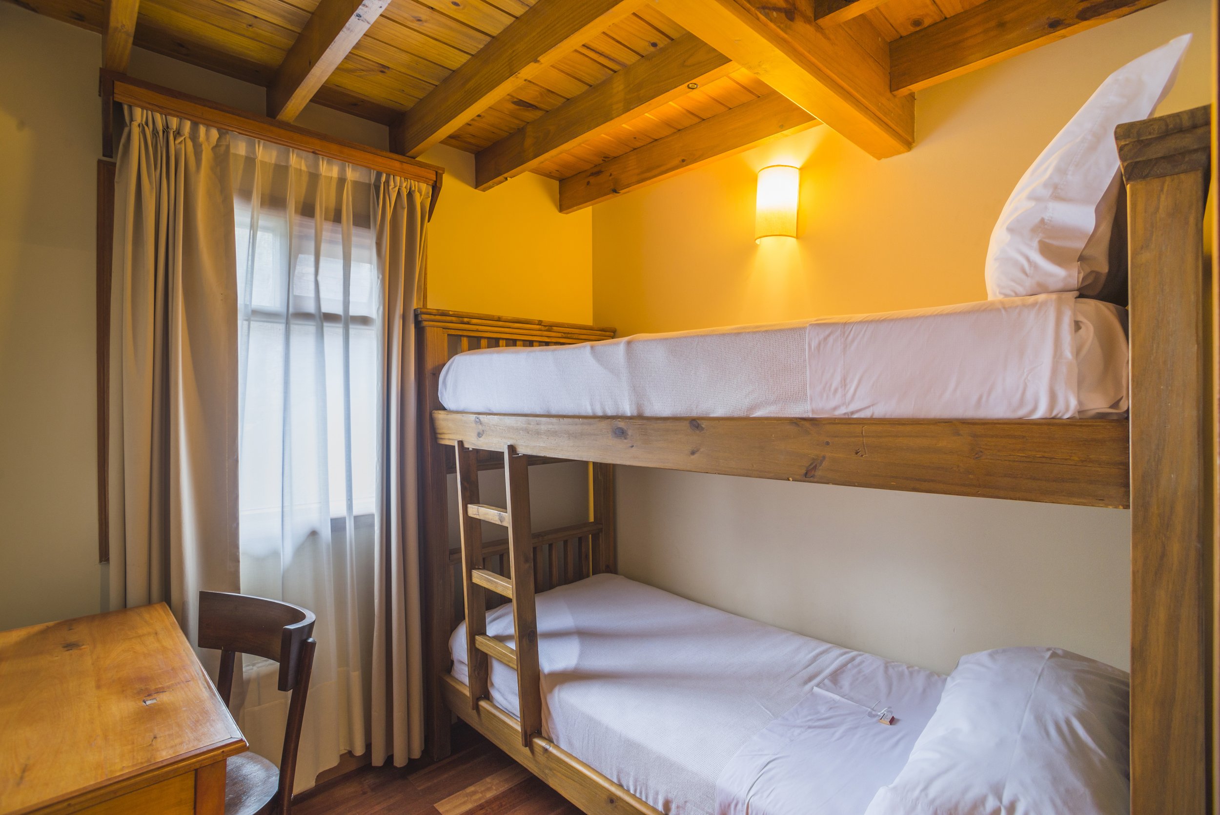 Habitación con cama cucheta de madera, techo con tirantes a la vista y ventana con cortinas.