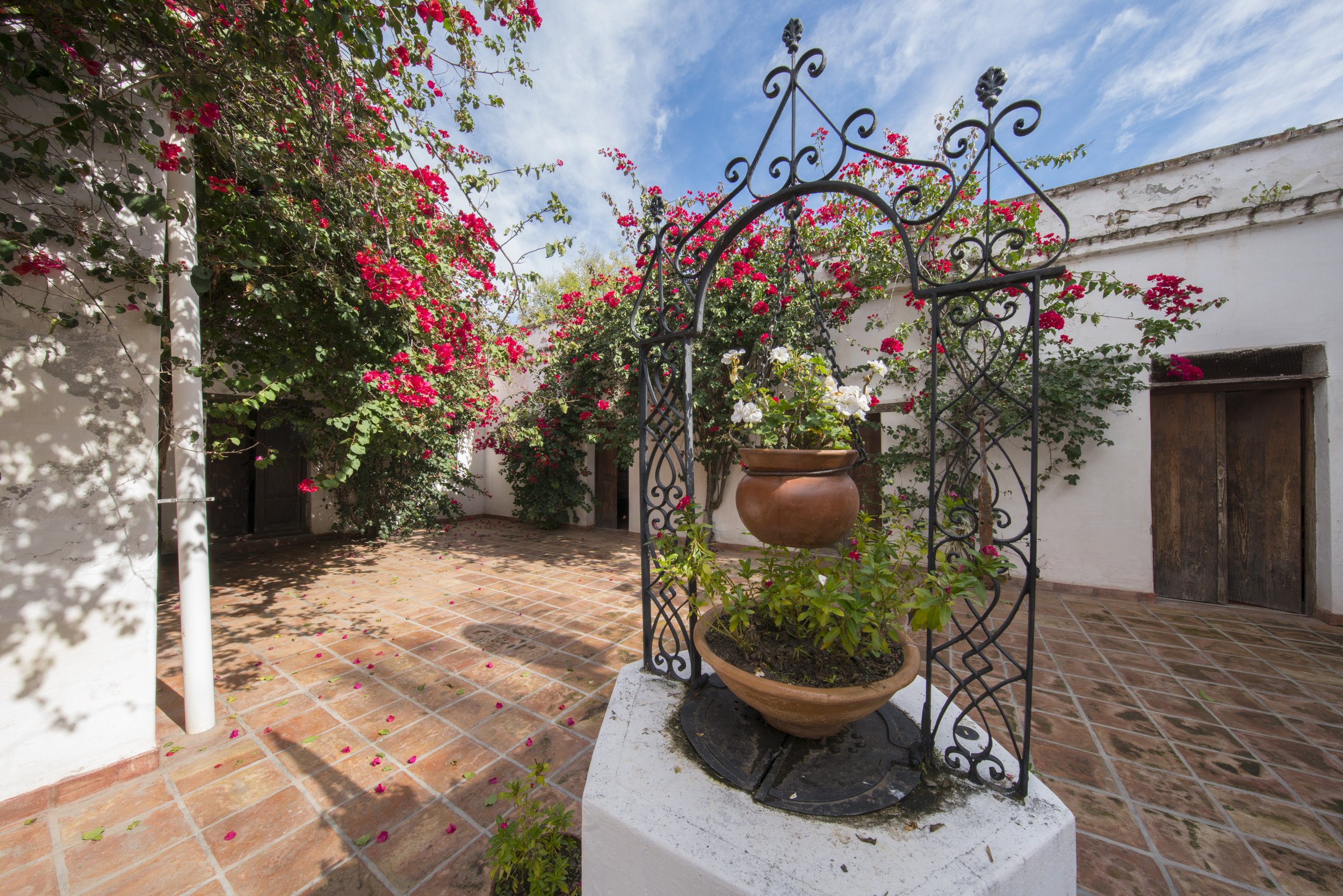 Patio colonial con piso de baldosas rojas, enredaderas con flores fucsias y un aljibe de hierro forjado.