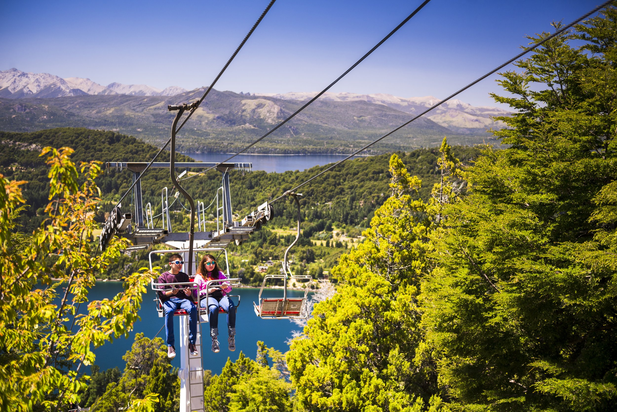 Dos personas ascienden en la aerosilla del Cerro Campanario con vista a los lagos y montañas de Bariloche.