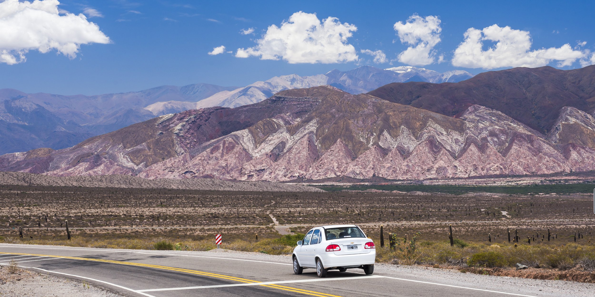 Auto blanco circulando por una ruta asfaltada frente a cerros coloridos en el Parque Nacional Los Cardones.
