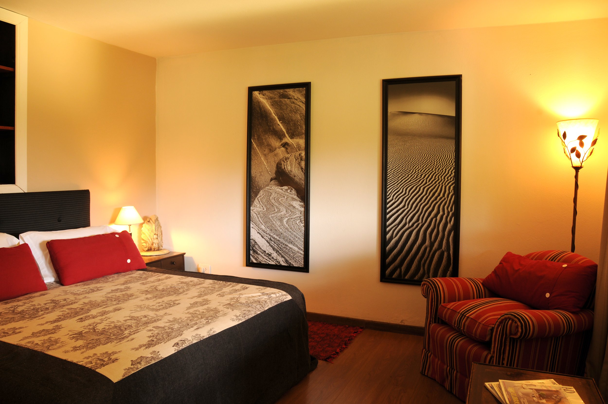 Habitación de hotel con cama matrimonial, sillón a rayas rojas, lámparas encendidas y cuadros en la pared.