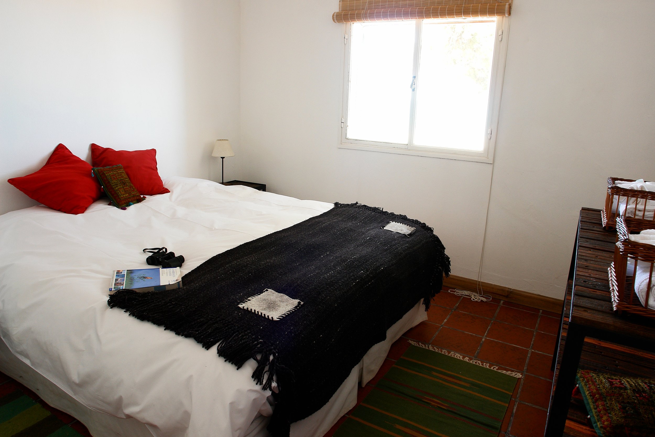 Habitación con cama doble, manta tejida negra, almohadones rojos, piso de baldosas y ventana iluminada por luz natural.
