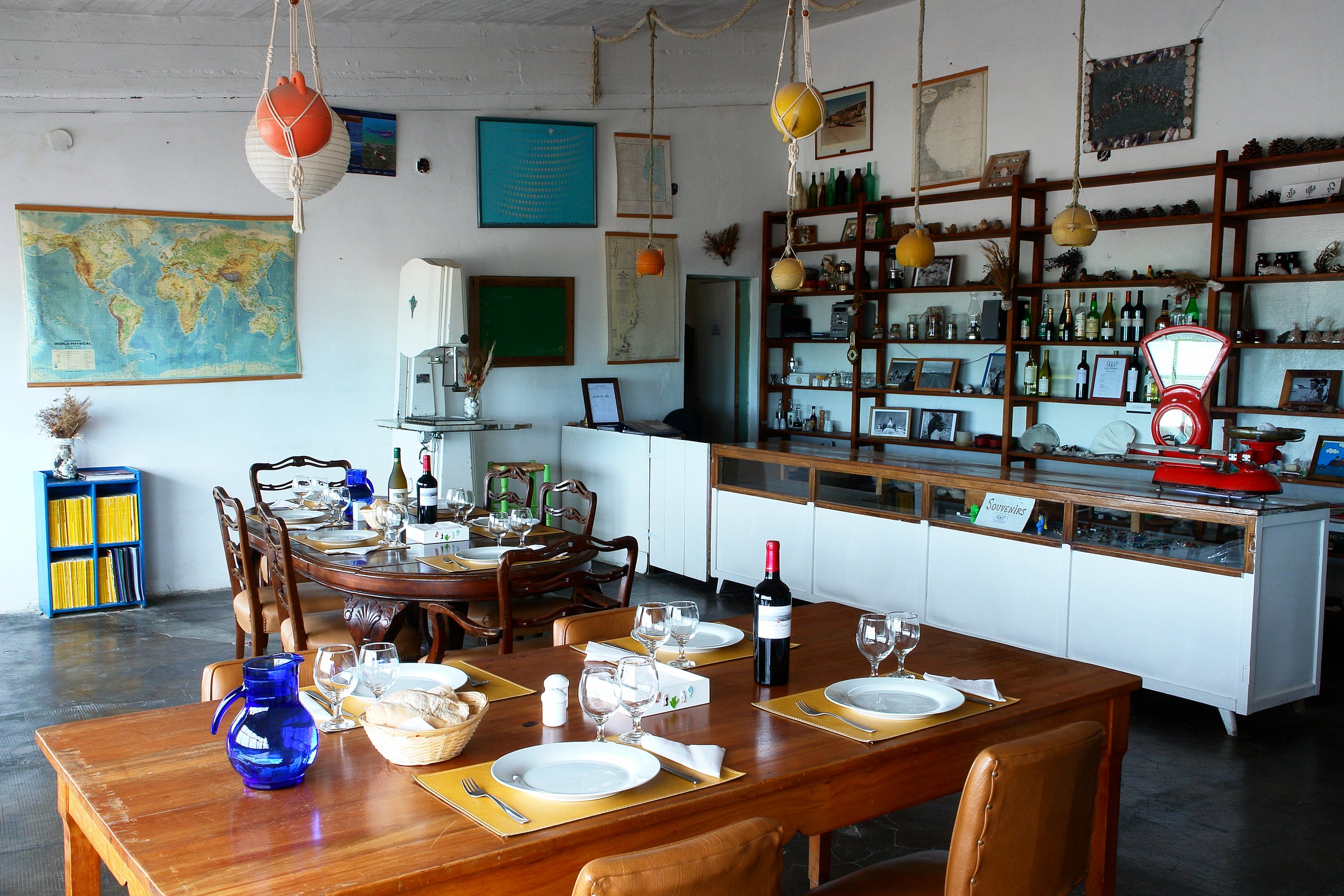 Comedor rústico con mesas de madera preparadas para el almuerzo y estanterías con objetos antiguos y botellas.