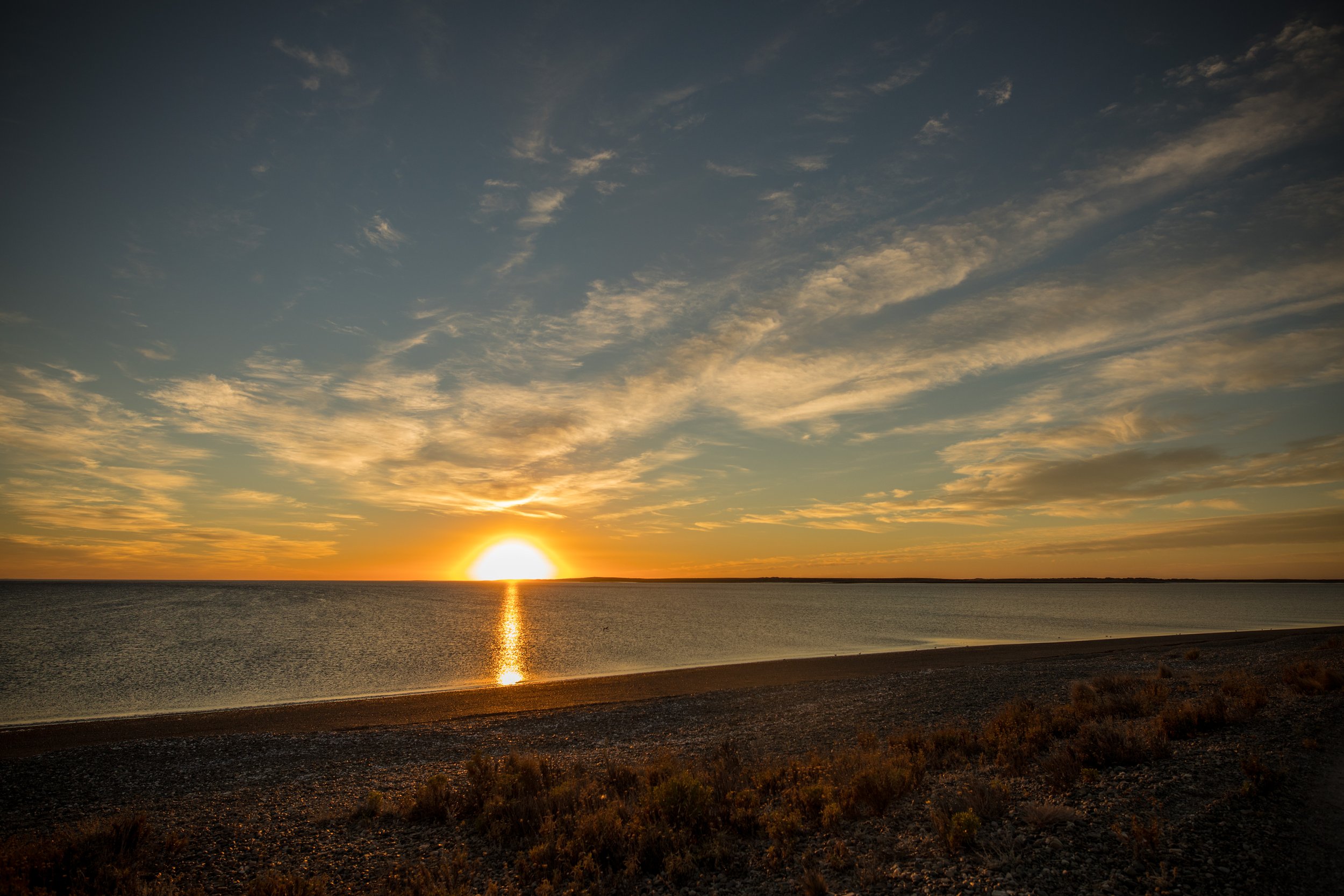 Amanecer sobre la costa marítima con el sol reflejado en el agua y cielo con nubes dispersas.