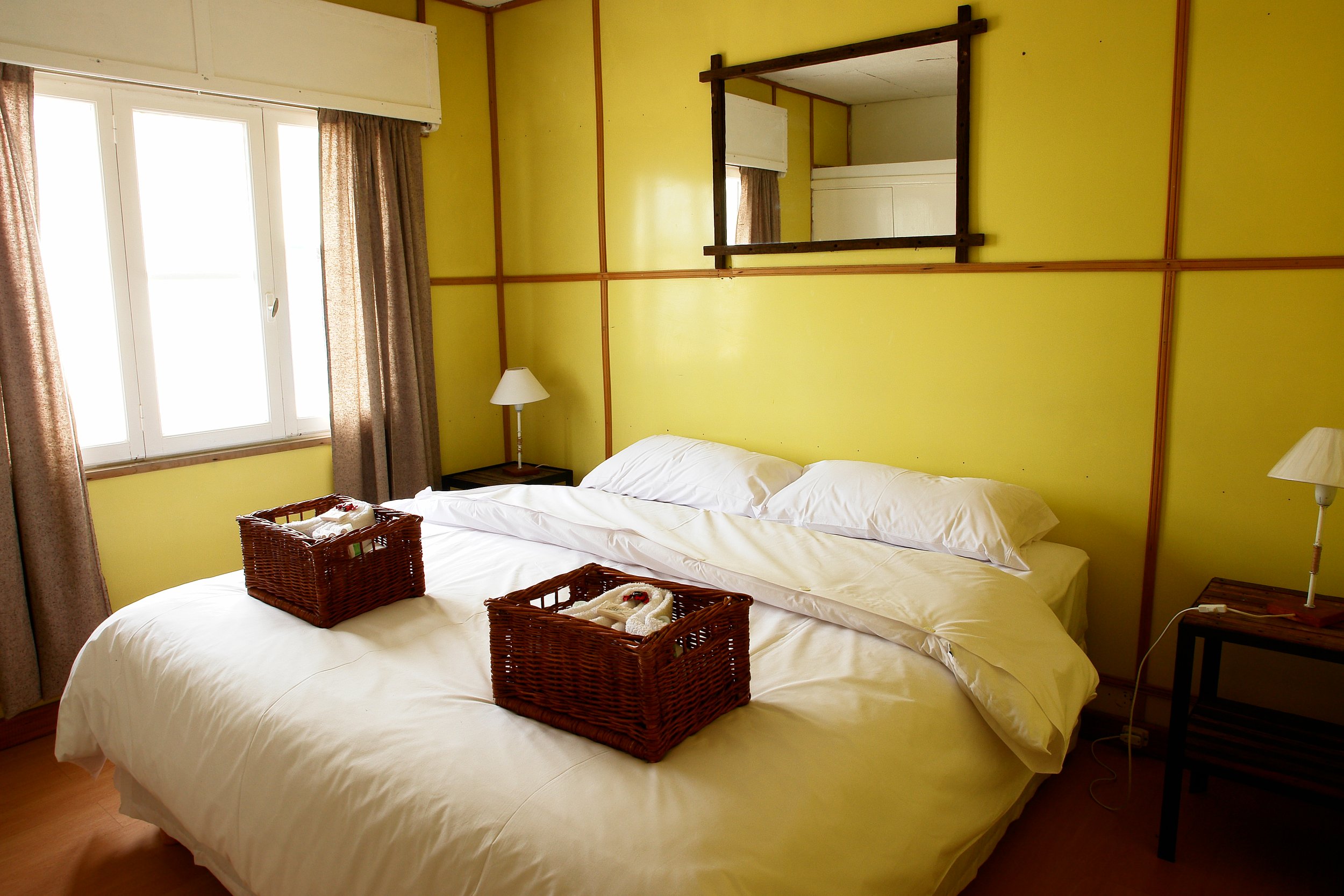 Habitación con paredes amarillas, cama matrimonial tendida de blanco con canastos de mimbre y ventana luminosa.