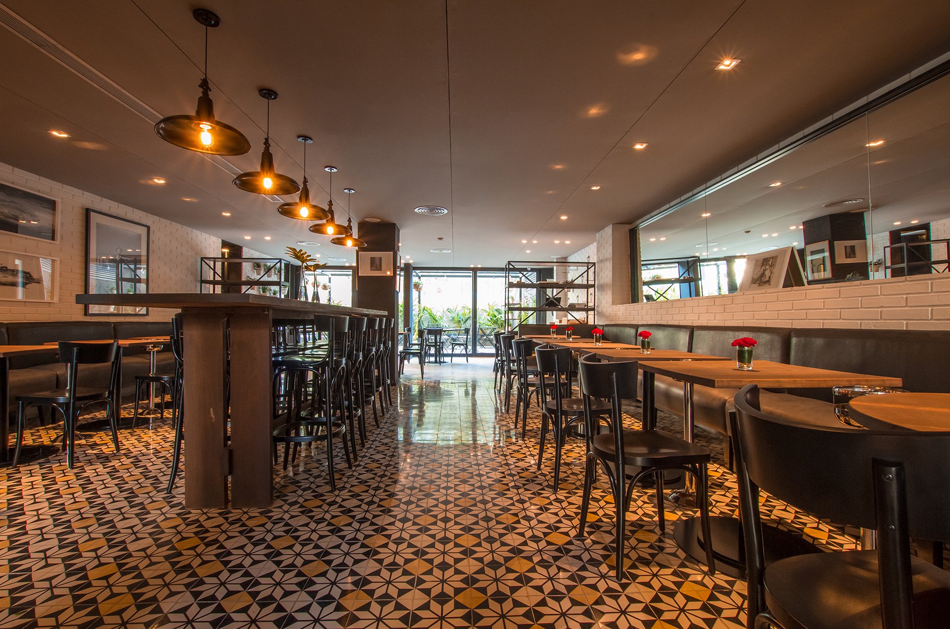 Salón comedor de hotel con mesas de madera, sillas negras, piso de mosaicos y lámparas colgantes.