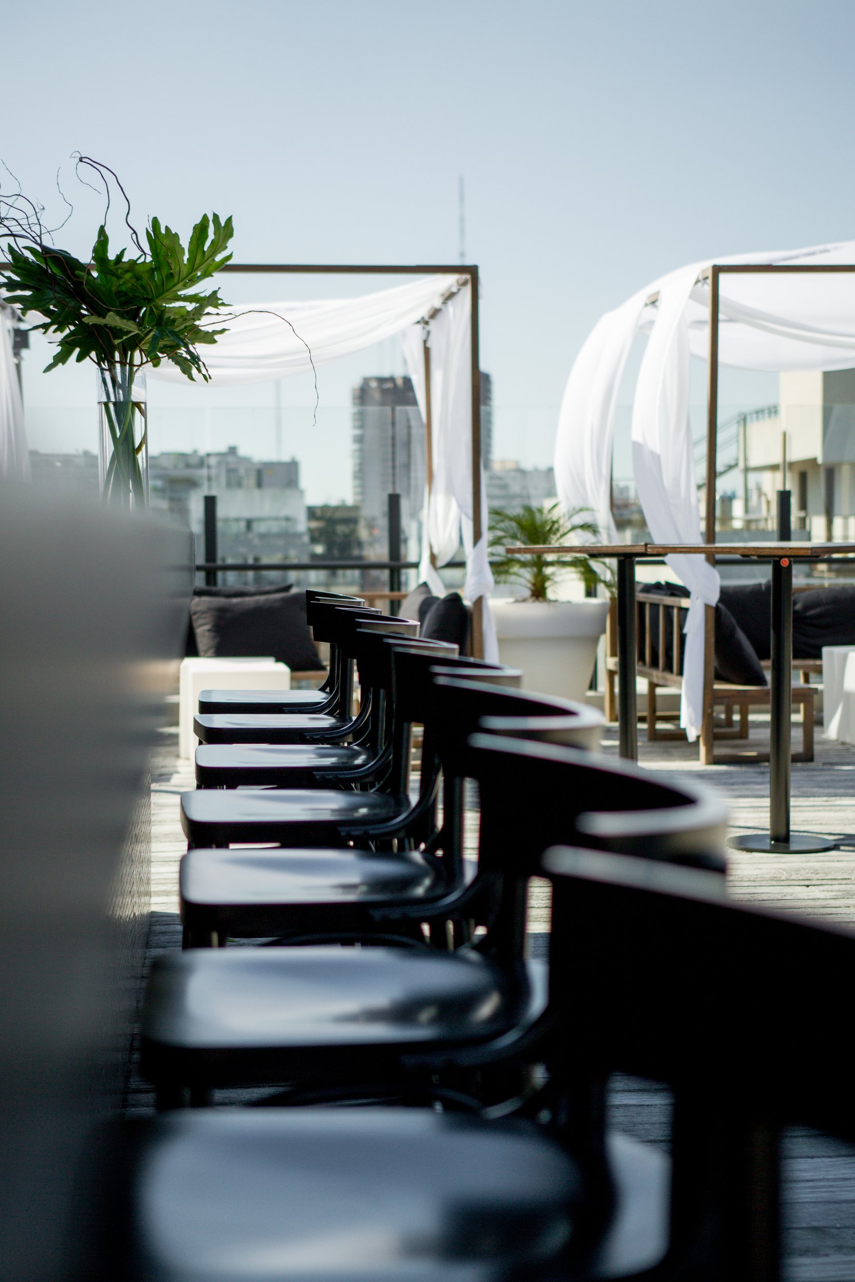 Terraza bar con banquetas negras alineadas, gazebo con telas blancas y vista a la ciudad.