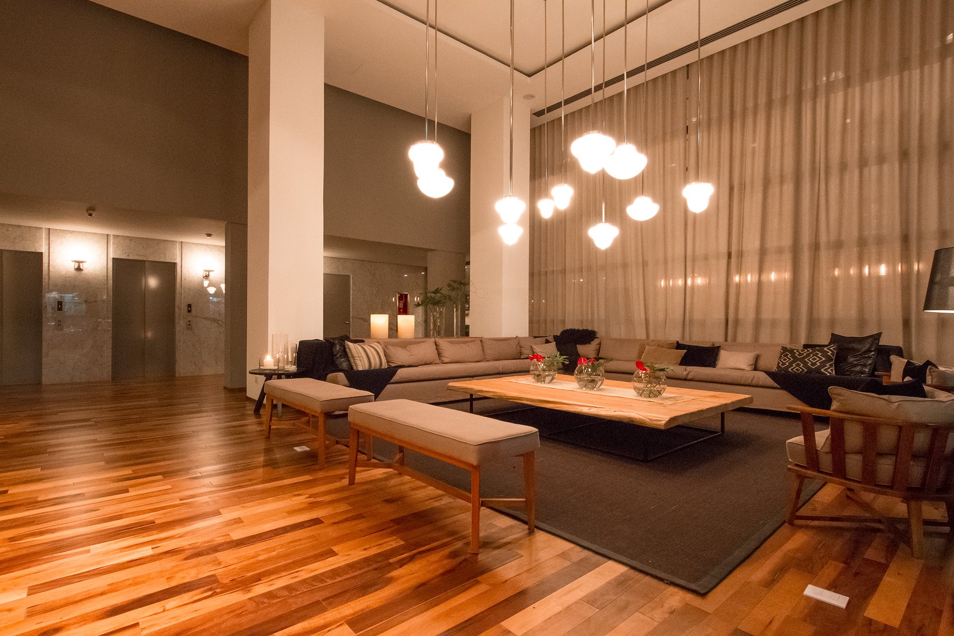 Lobby de hotel moderno con sofás amplios, piso de madera, mesas bajas y lámparas colgantes encendidas.