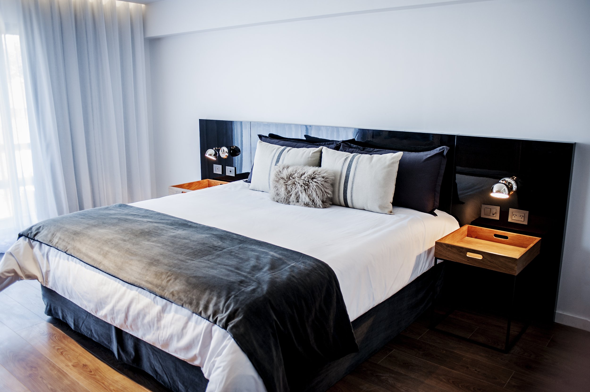 Habitación de hotel con cama doble, respaldo negro brillante, mesas de luz de madera y cortinas blancas.