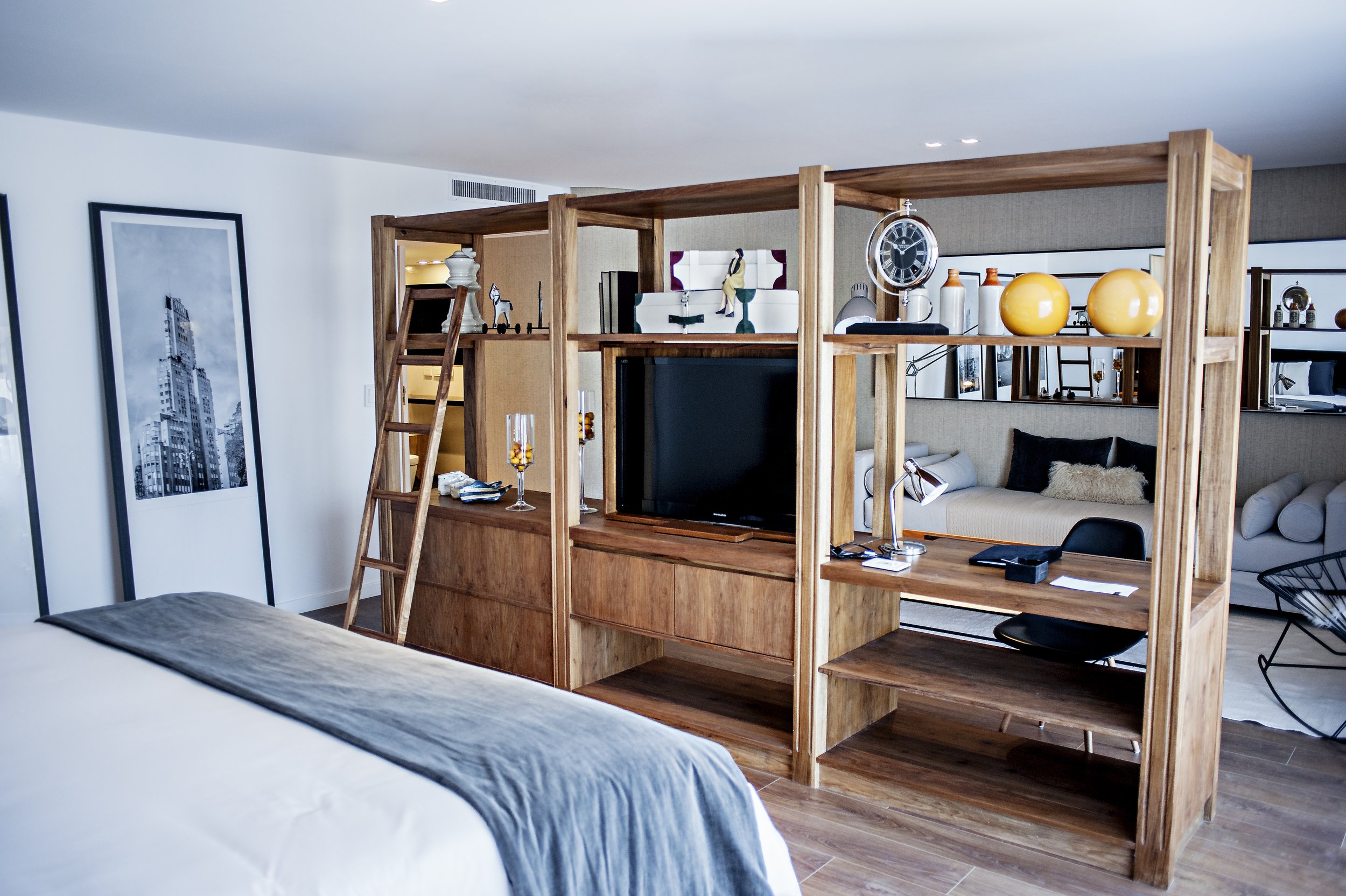Habitación de hotel con cama en primer plano, estantería divisoria de madera, escritorio y zona de estar.