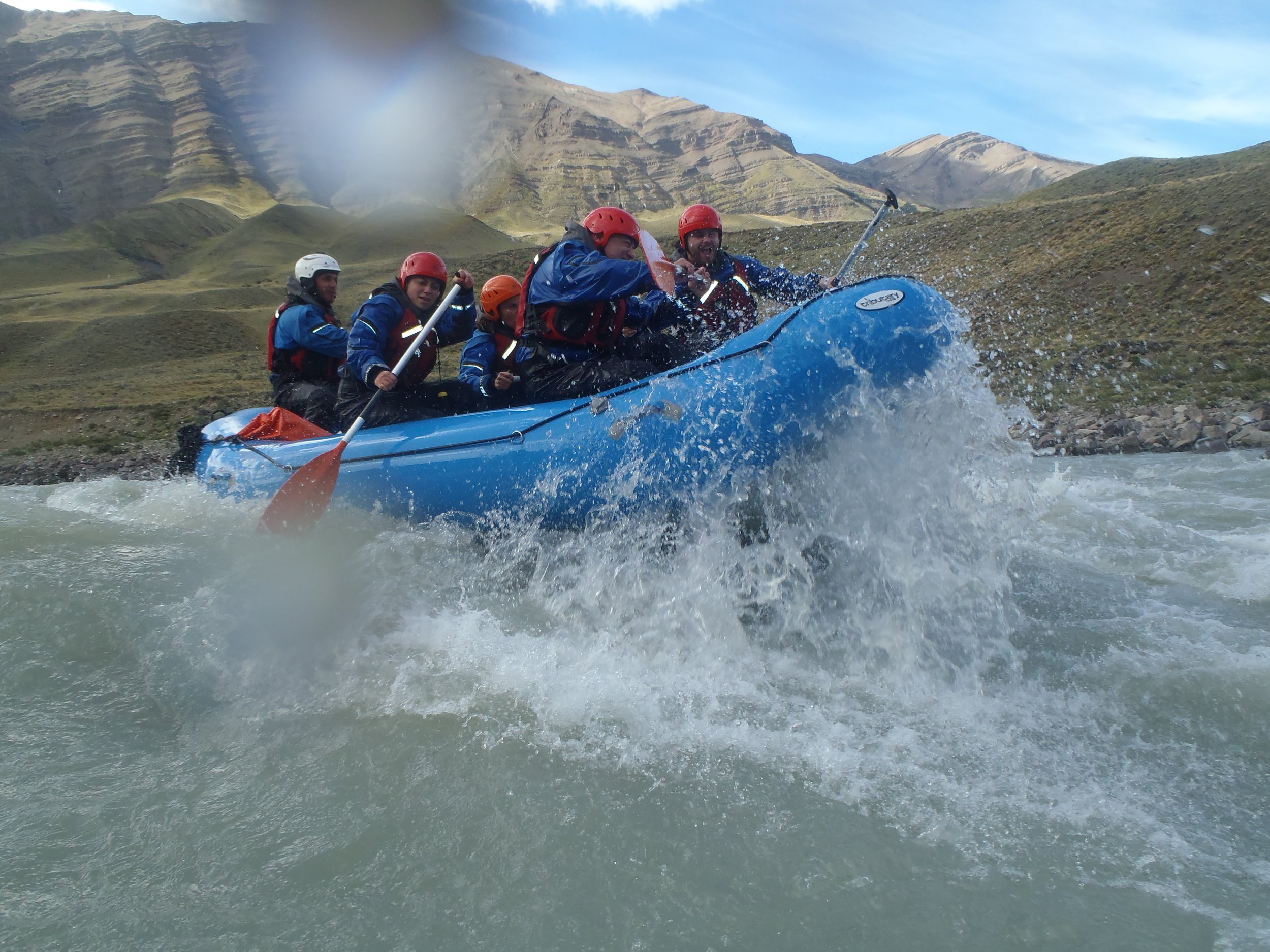 Grupo de personas practicando rafting en un gomón azul sobre los rápidos de un río de montaña.
