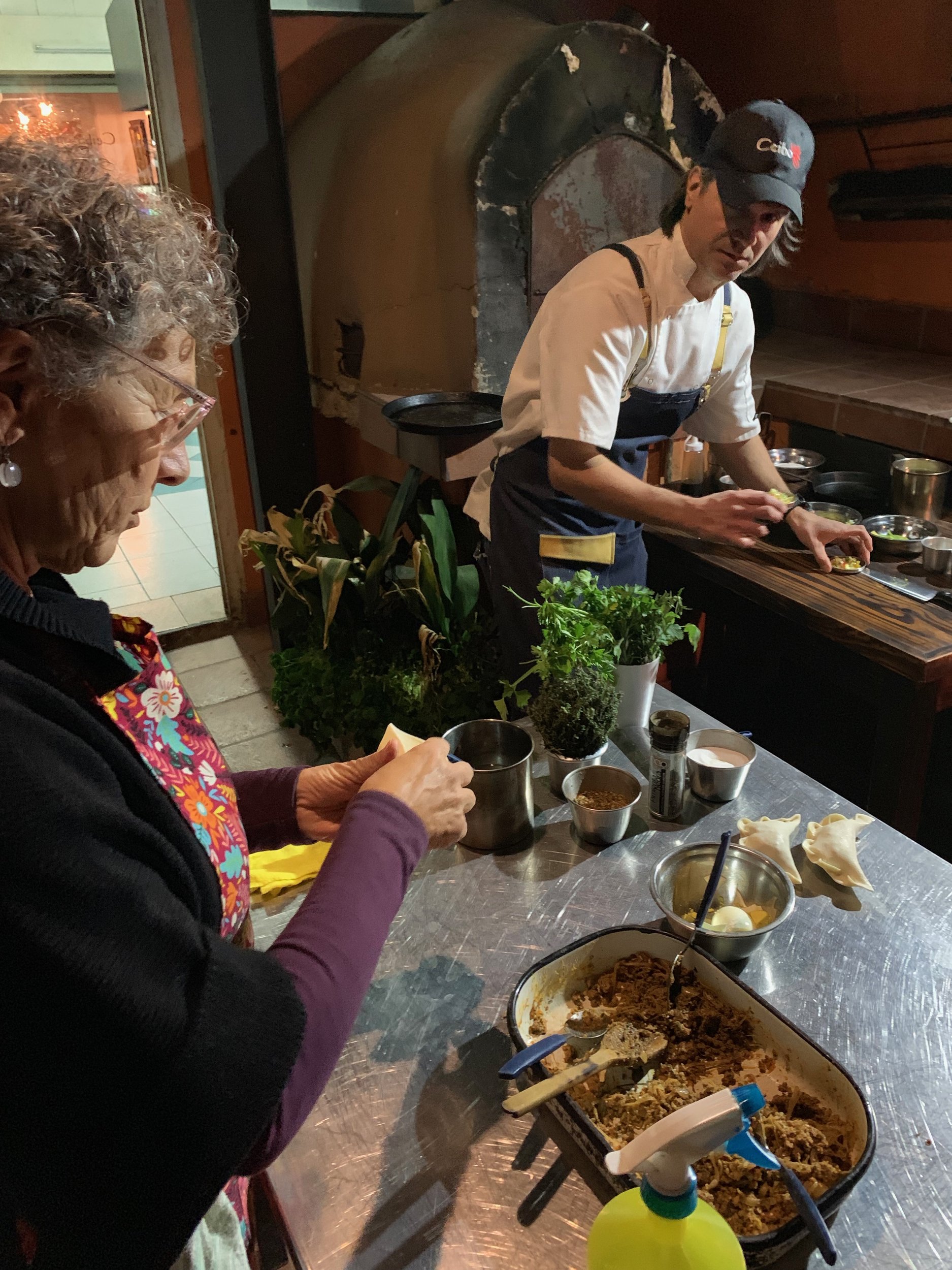 Una mujer y un cocinero preparando empanadas durante una clase de cocina cerca de un horno de barro.