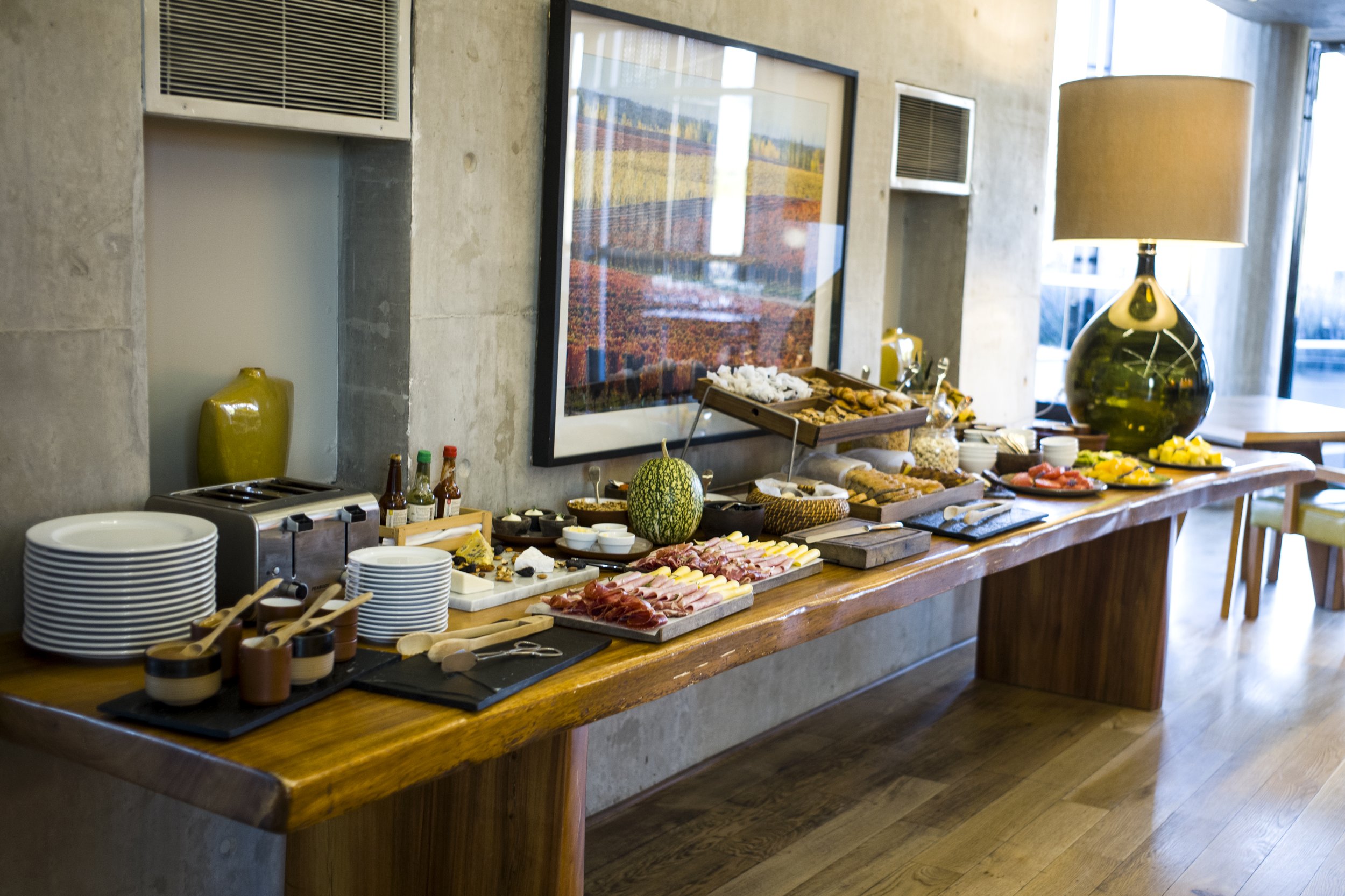 Mesa de madera con variedad de fiambres, frutas, panes y vajilla en el buffet de un hotel.