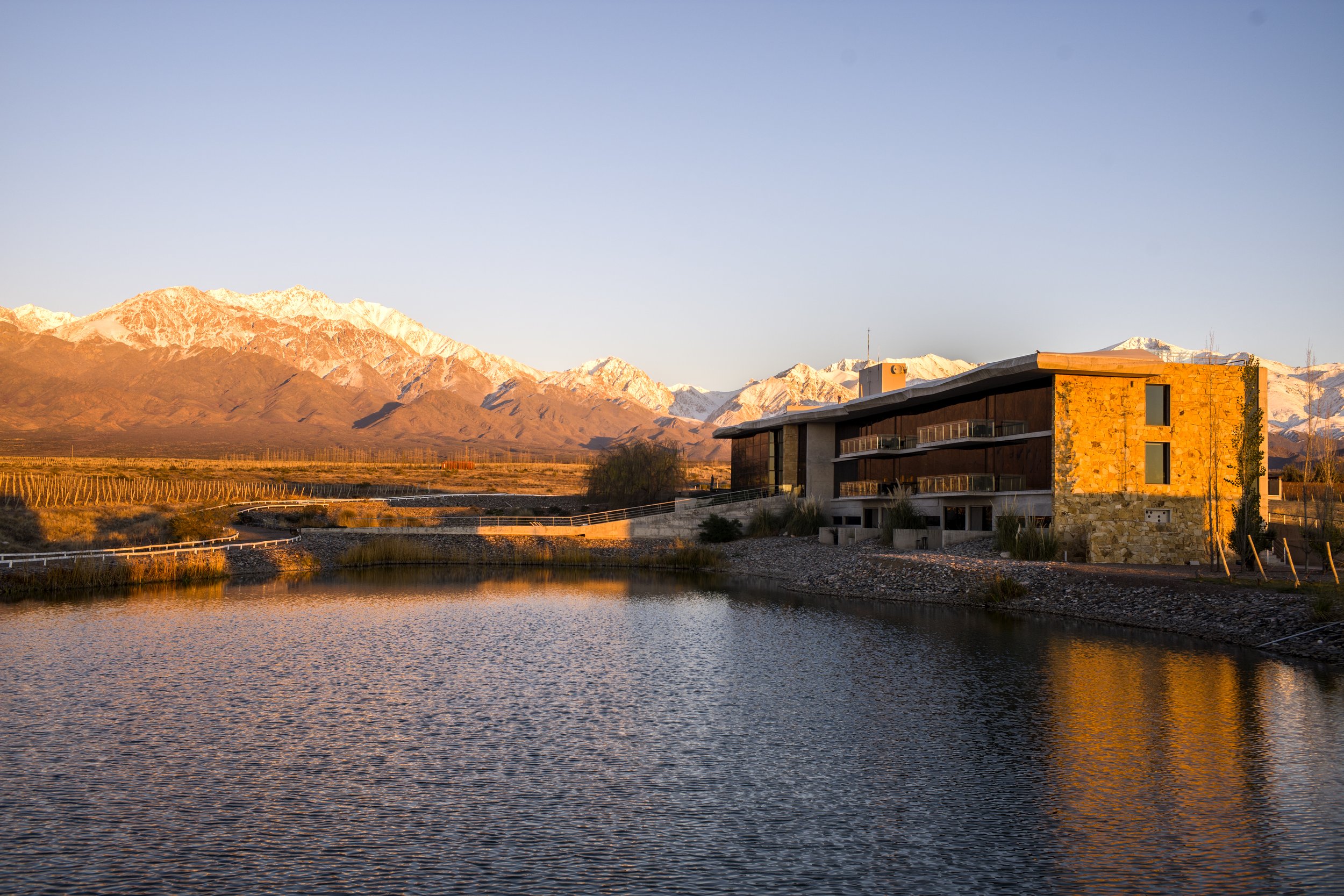 Edificio moderno de hotel junto a una laguna con viñedos y la cordillera nevada de fondo.