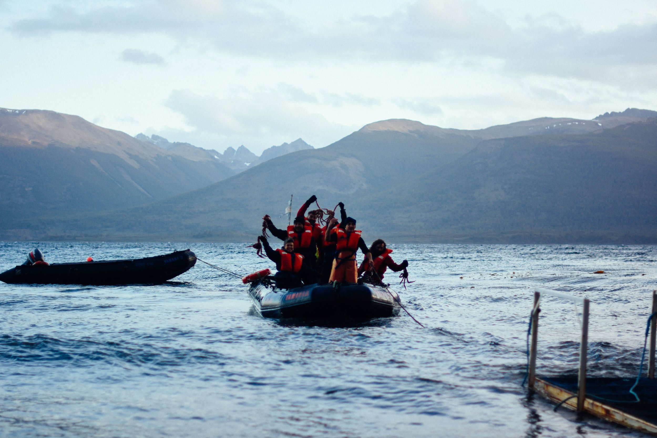 Grupo de personas con chalecos salvavidas en un gomón sosteniendo centollas en el Canal Beagle con montañas de fondo.