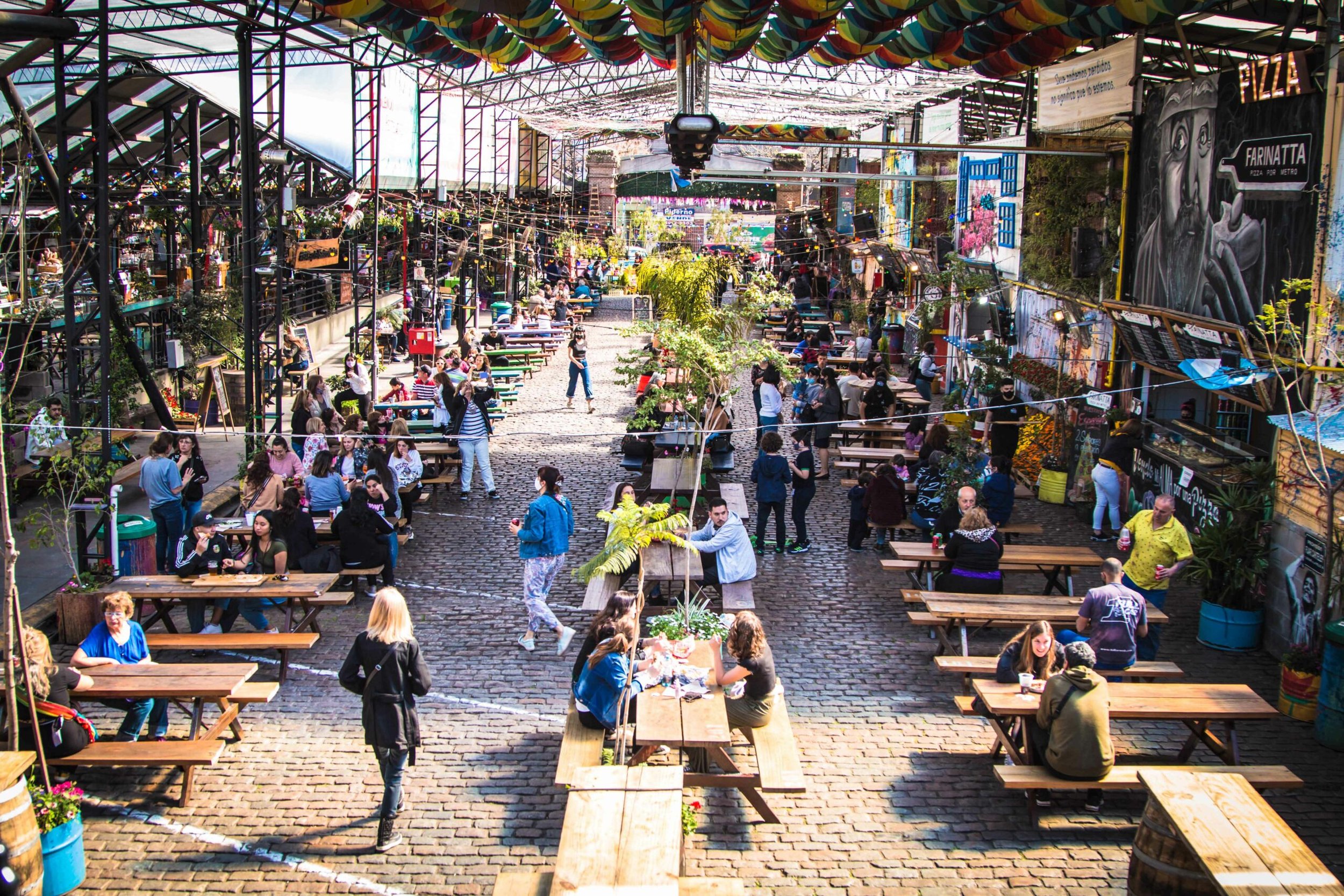 Personas sentadas en mesas comunitarias al aire libre en un patio gastronómico y cultural adoquinado.