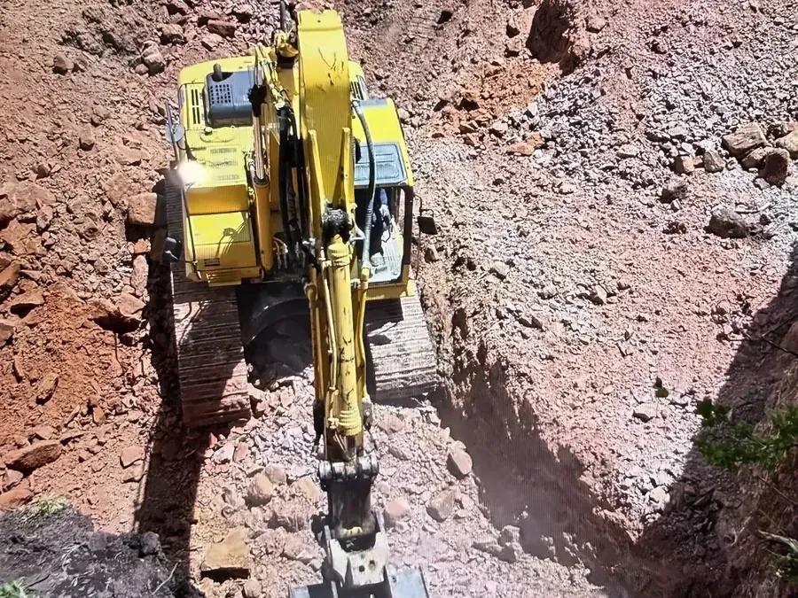 Excavator digging a deep cut through red rock and soil on a wooded mountain lot