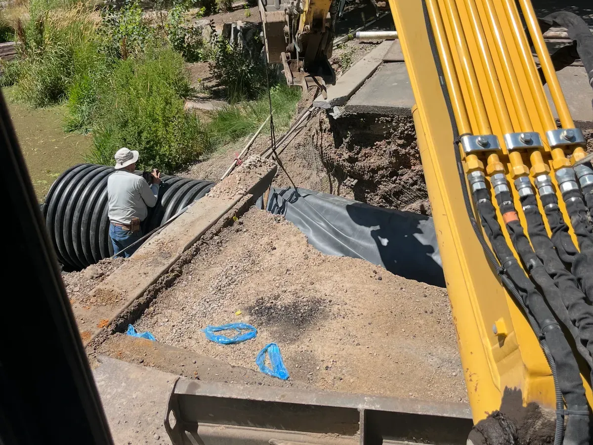 Crew setting a large corrugated drainage culvert in a lined trench beside an excavator