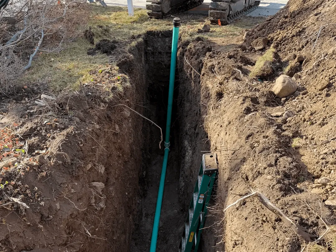 New green sewer pipe set in a deep trench running from a house toward the street, with a mini excavator beyond