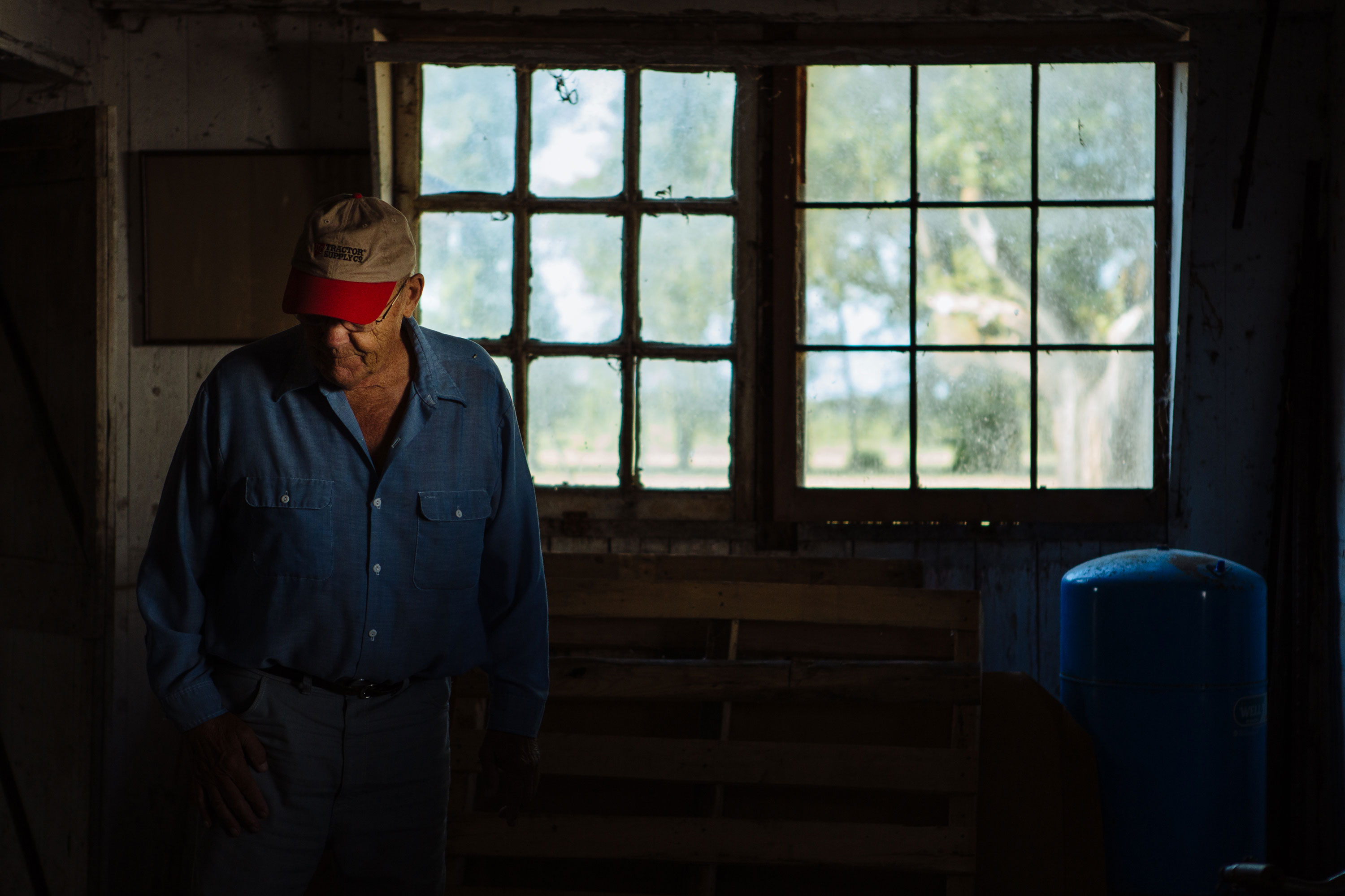 Elderly man in a blue shirt and beige cap standing inside a dimly lit rustic room with a wooden pallet and blue tank near a window showing an outdoor view.