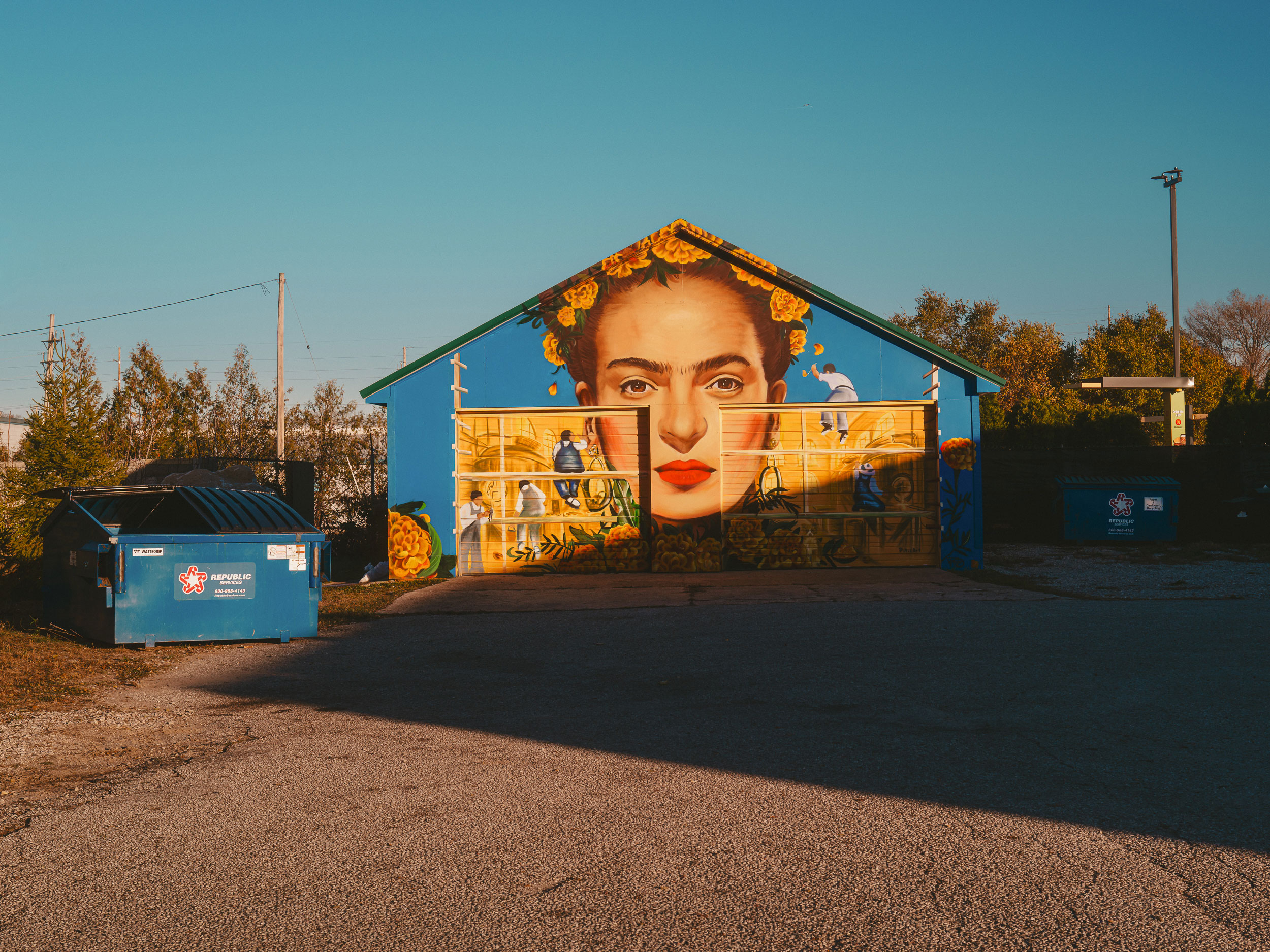 A large mural painted on a blue building features a close-up of a woman's face with red lipstick and yellow flowers in her hair, bordered by smaller depictions of people engaging in various activities on a yellow background that covers the doors and lower walls.
