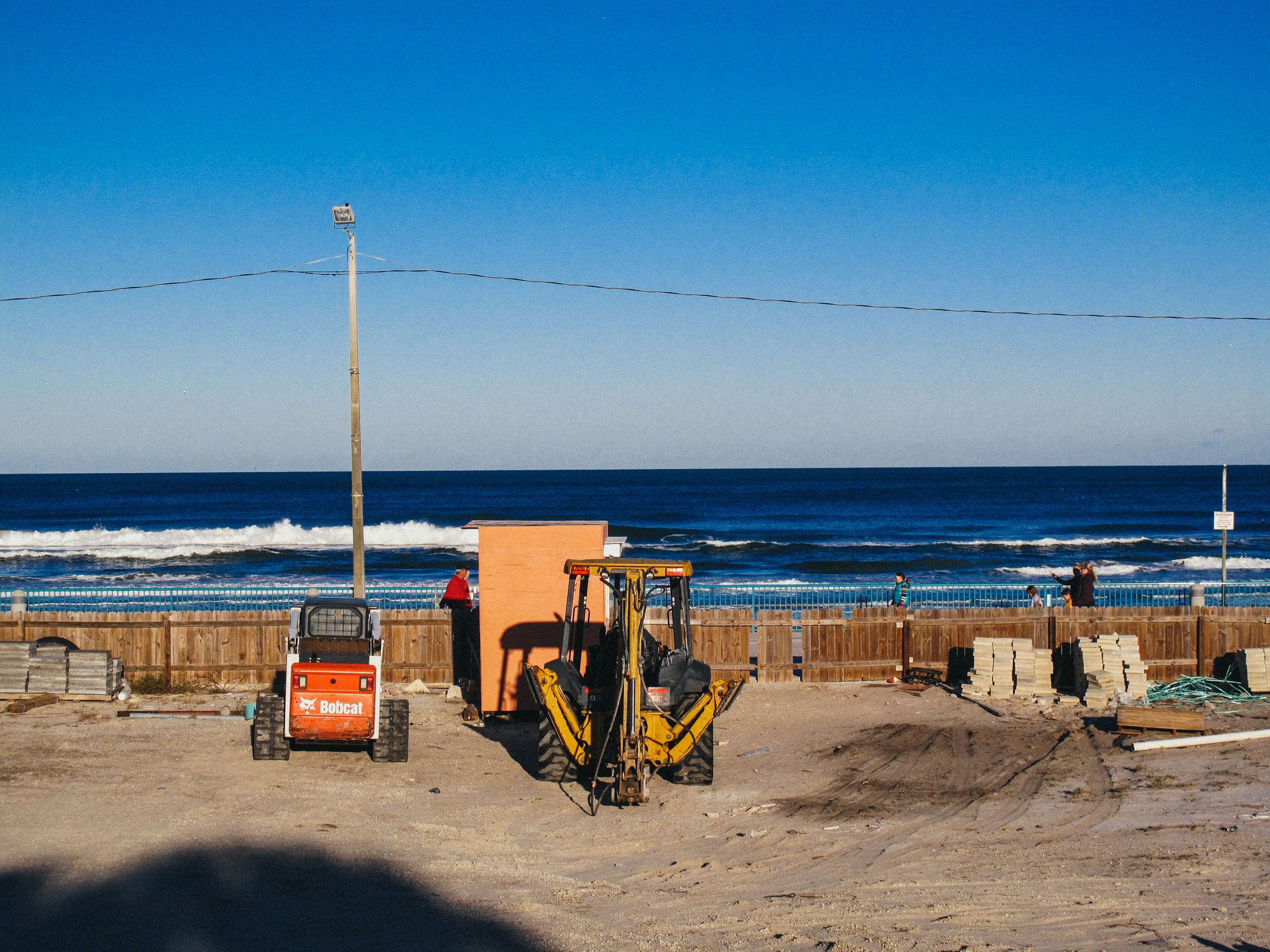 Construction site near a beach with a Bobcat loader and a yellow backhoe loader parked on sandy ground, a wooden fence separating the site from the ocean with waves, clear blue sky, and a few people walking or standing near the fence.