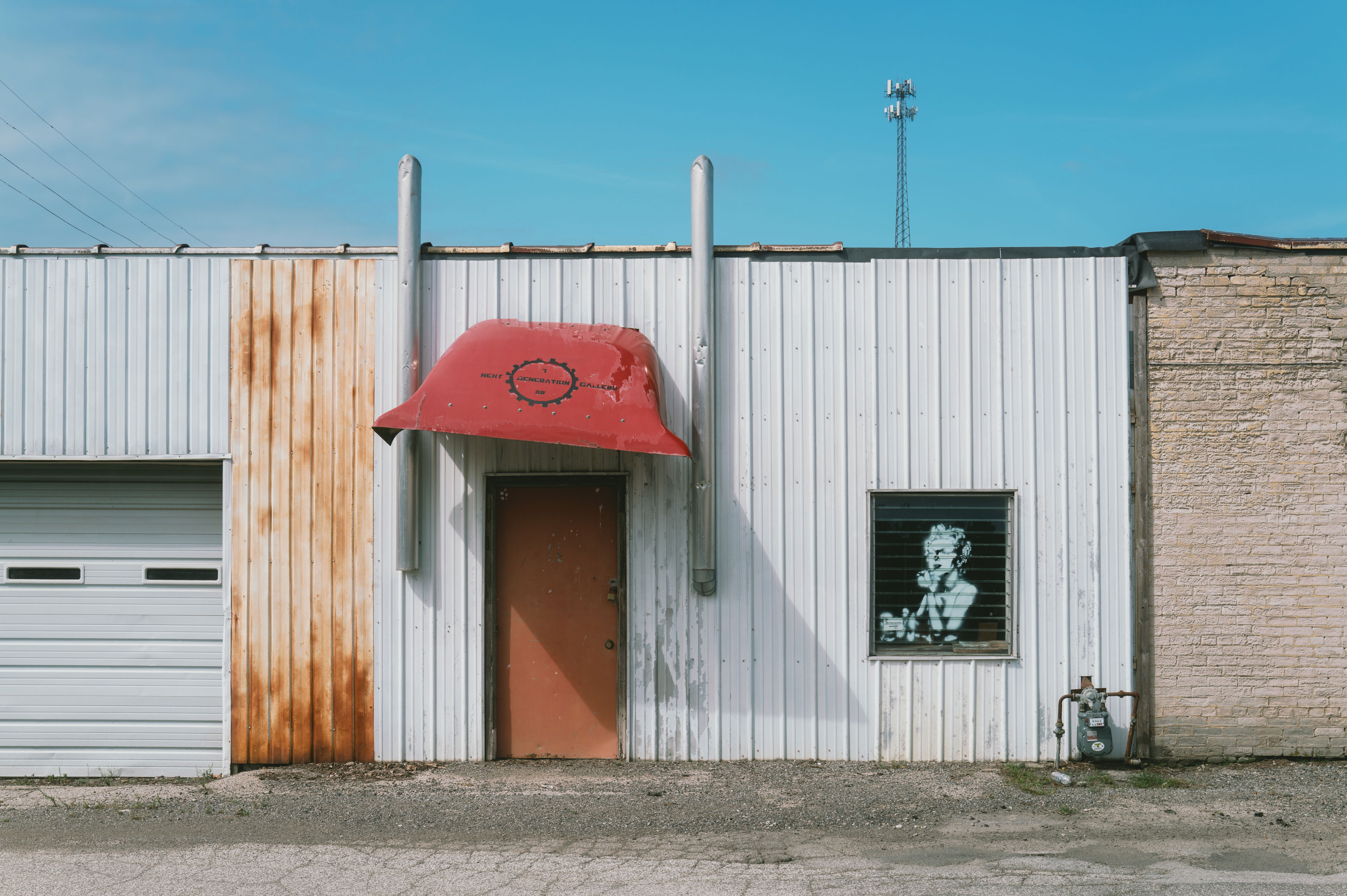 Exterior of a white metal-paneled building with rust stains on one section, featuring a red door under a red awning labeled 'Next Generation 08 Gallery' and a window with a black-and-white image of a woman, set against a clear blue sky with a tall communication tower in the background.